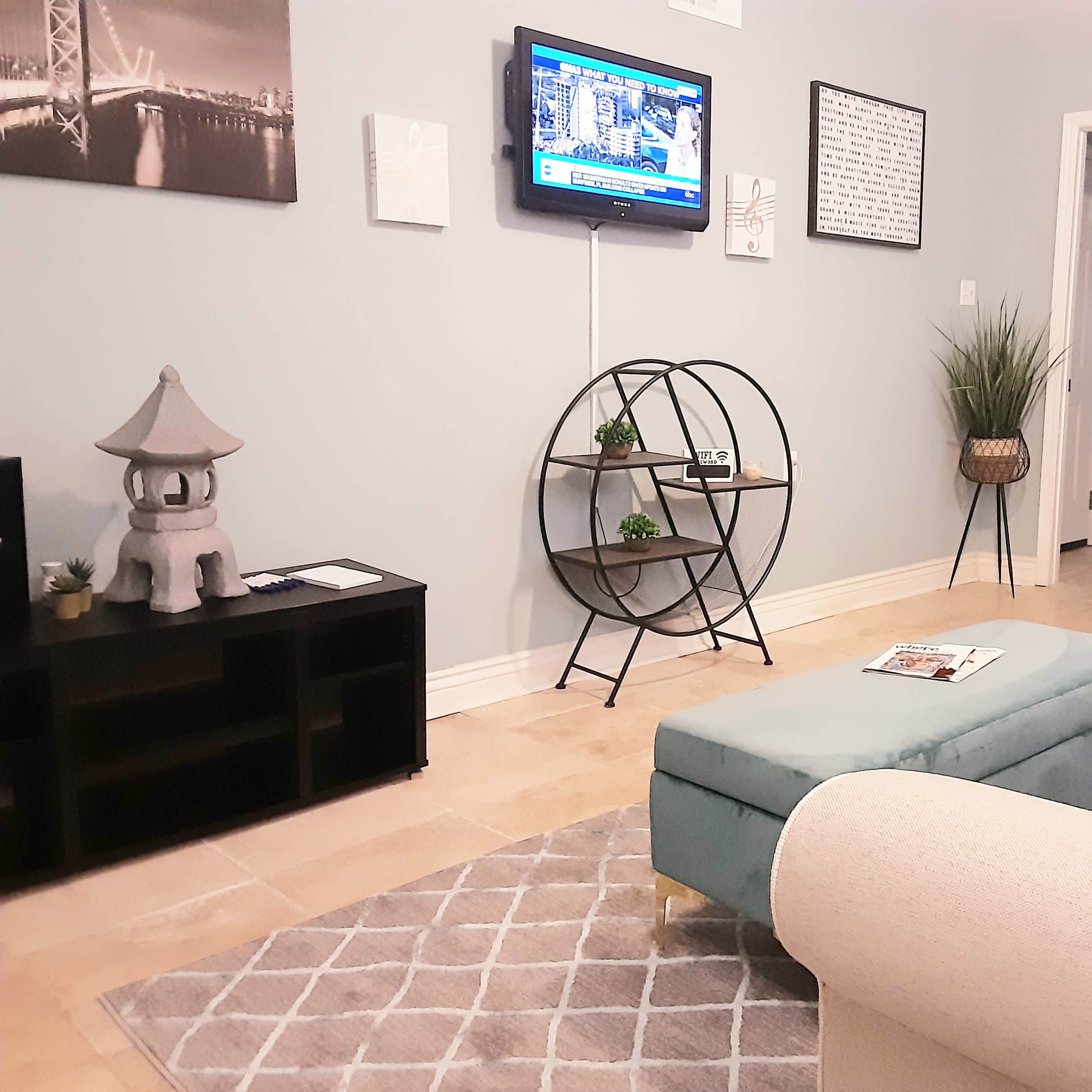 A modern living room with a gray sofa, an ottoman, a circular shelf displaying decorative items, and a wall-mounted television showing news.