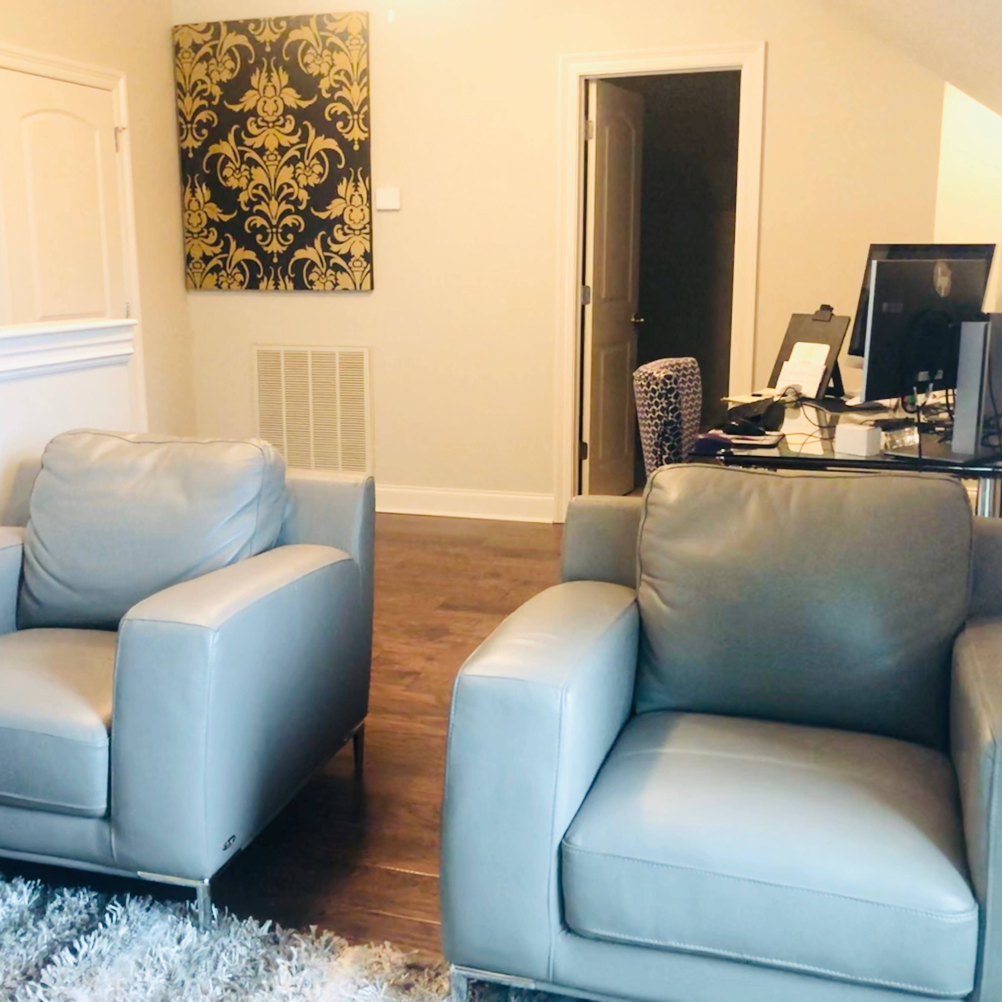 A room with two gray armchairs facing each other, a decorative wall hanging, and a desk with a computer in the background.