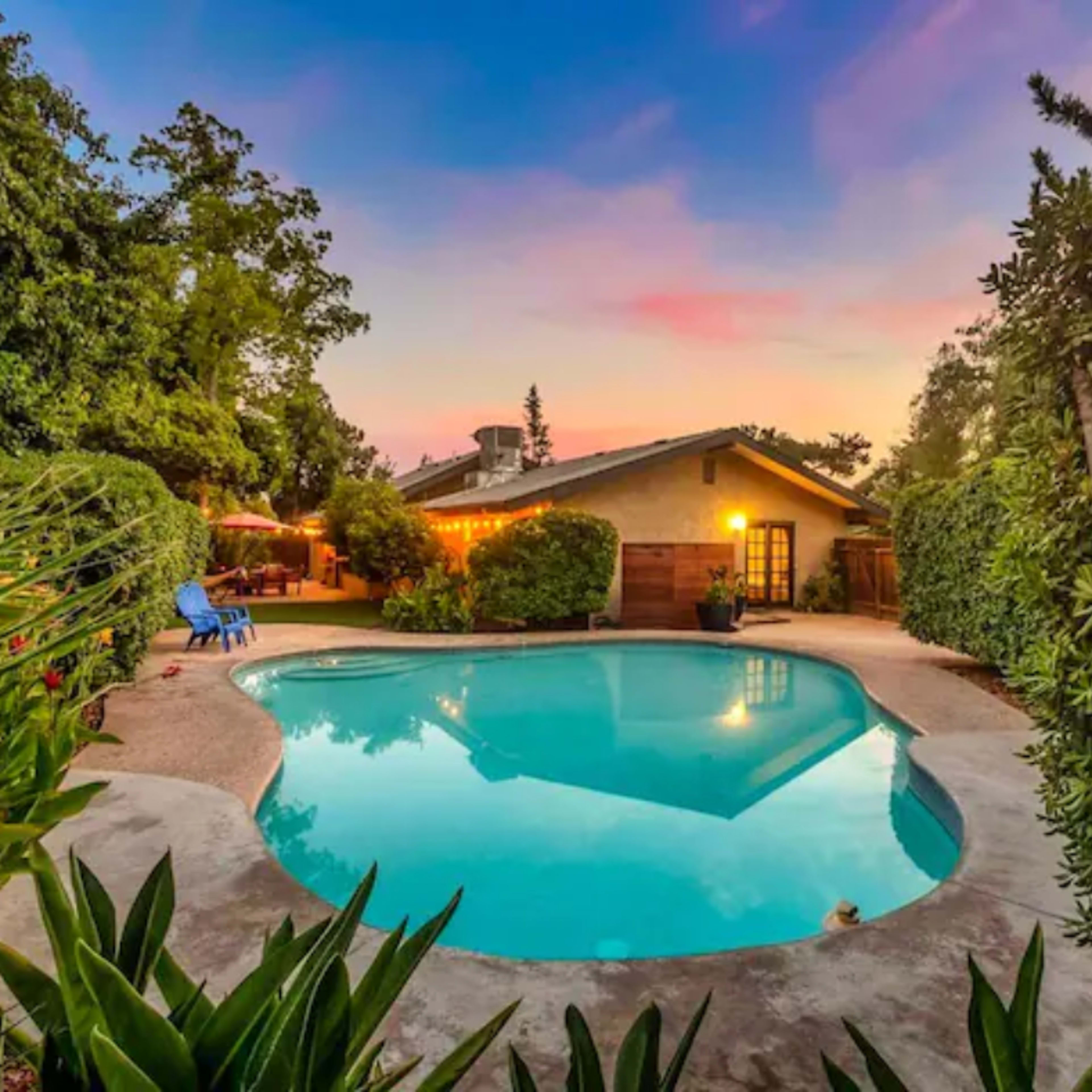A swimming pool is surrounded by greenery and well-maintained landscaping, with a house and patio visible in the background.