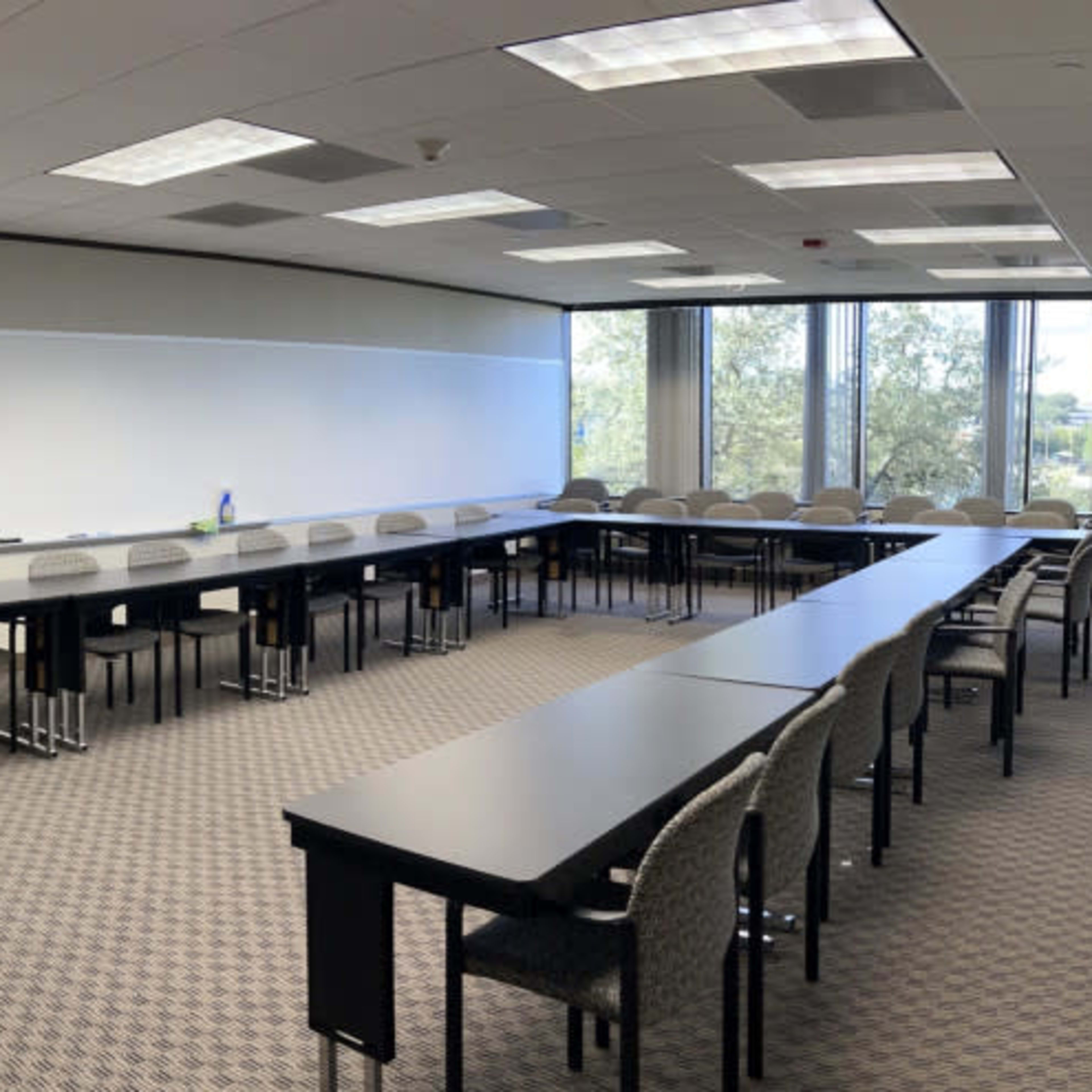 A conference room with multiple long tables arranged in a U-shape and chairs along the sides, surrounded by large windows.