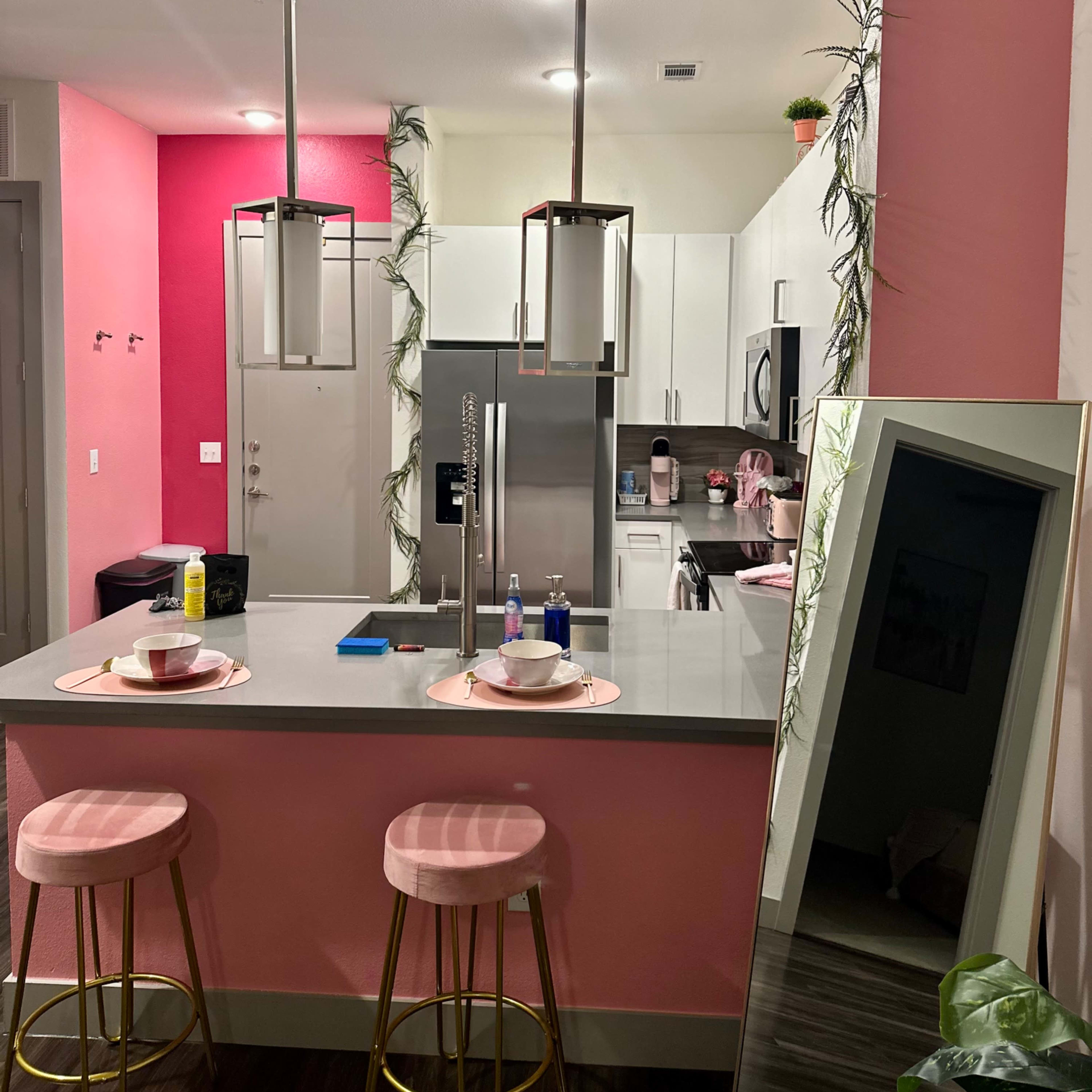 A modern kitchen area with pink accent walls, bar stools, and a mirror reflecting the space.