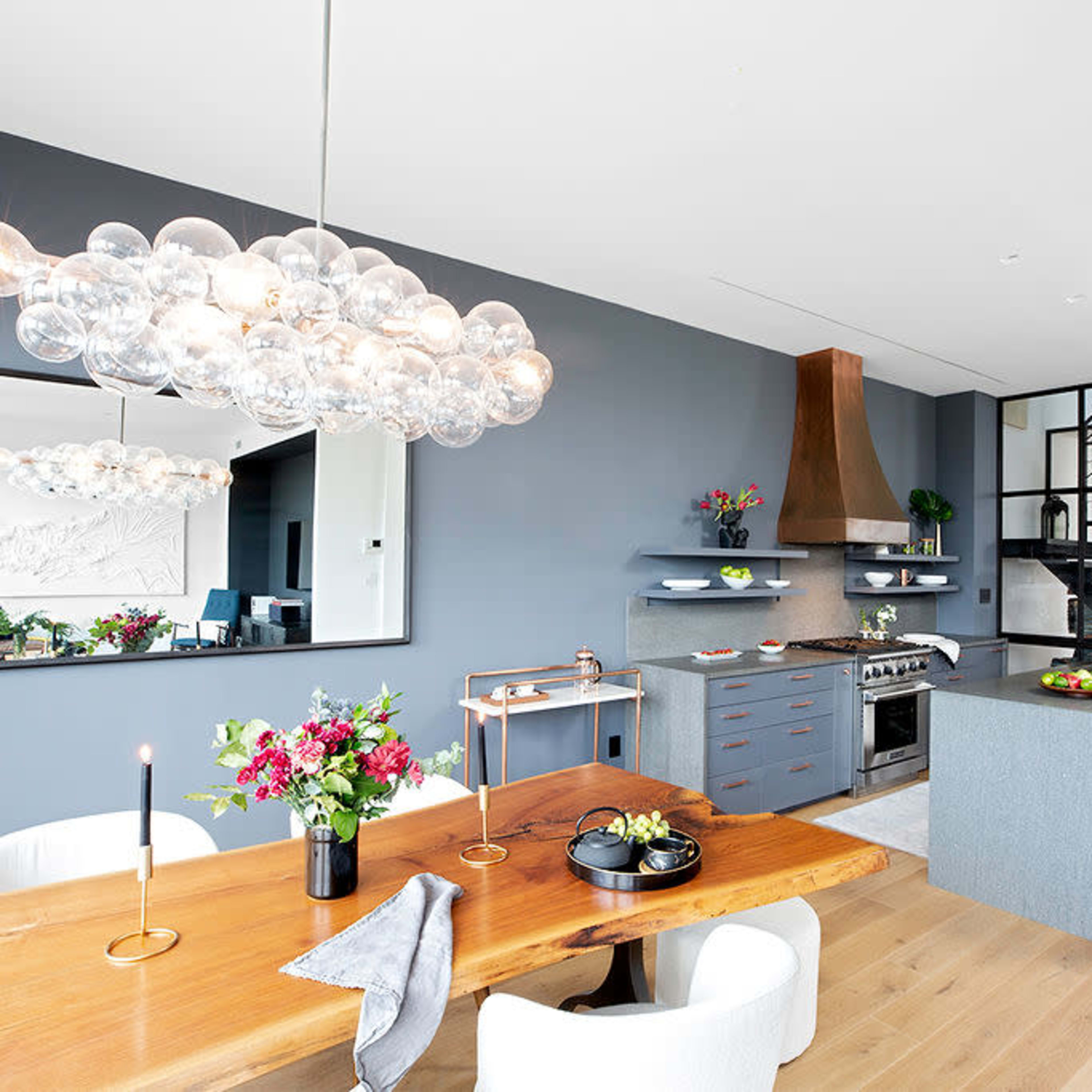 A modern kitchen and dining area with a large wooden table, sleek cabinetry, and a unique light fixture, complemented by a staircase in the background.