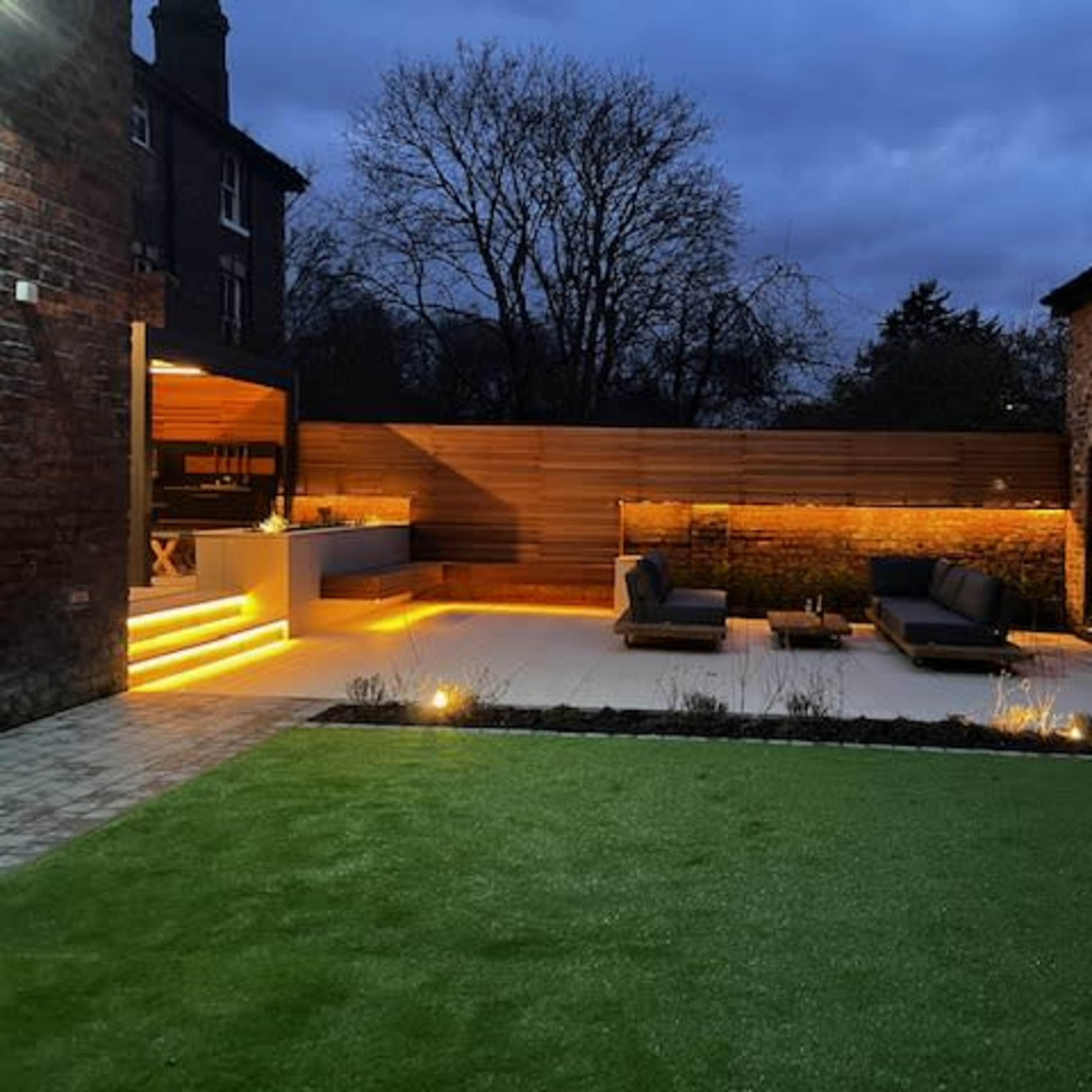 A modern outdoor patio with seating and landscape lighting, surrounded by brick walls and a grassy area under a twilight sky.