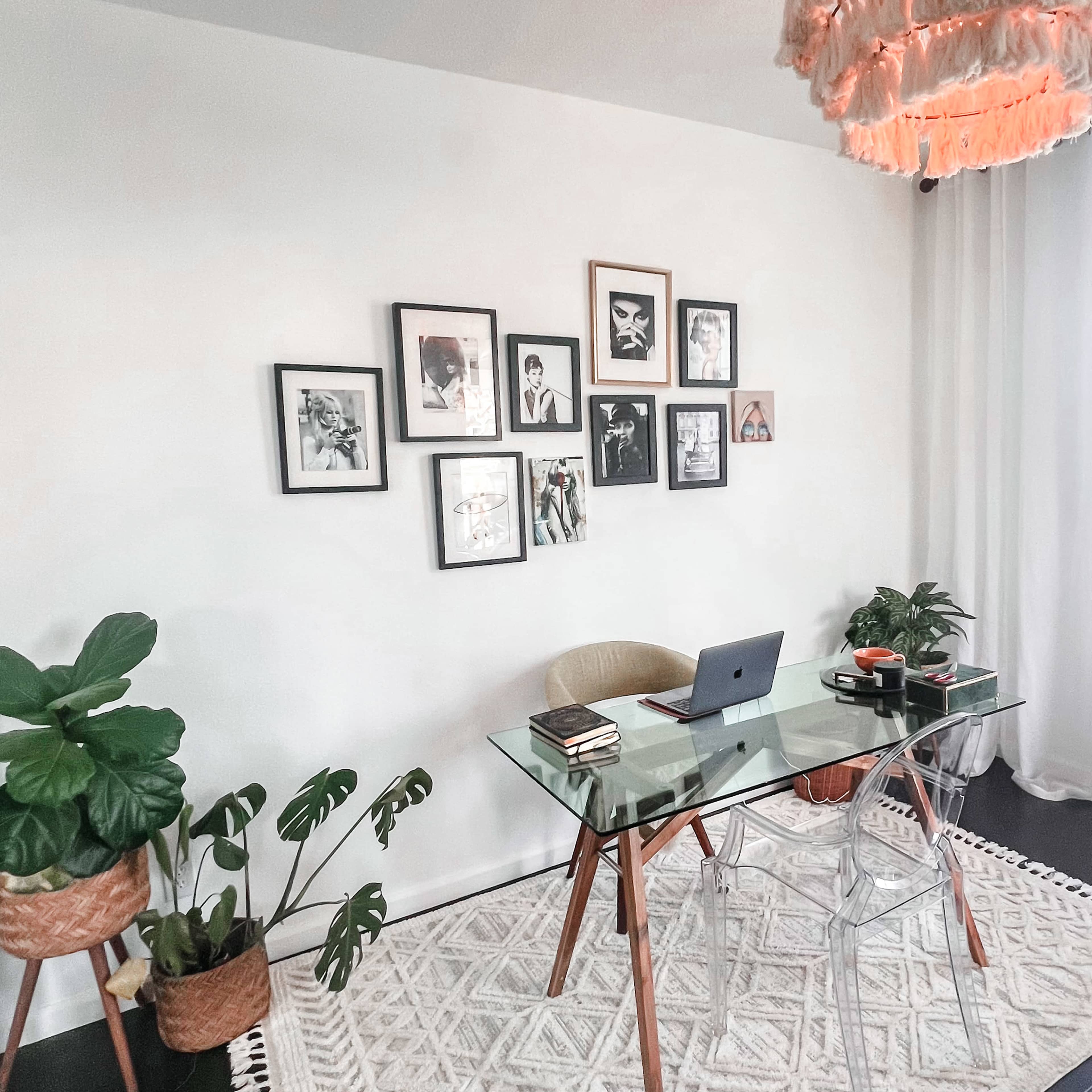 A modern workspace features a glass desk with a laptop, surrounded by framed photographs on the wall and potted plants.