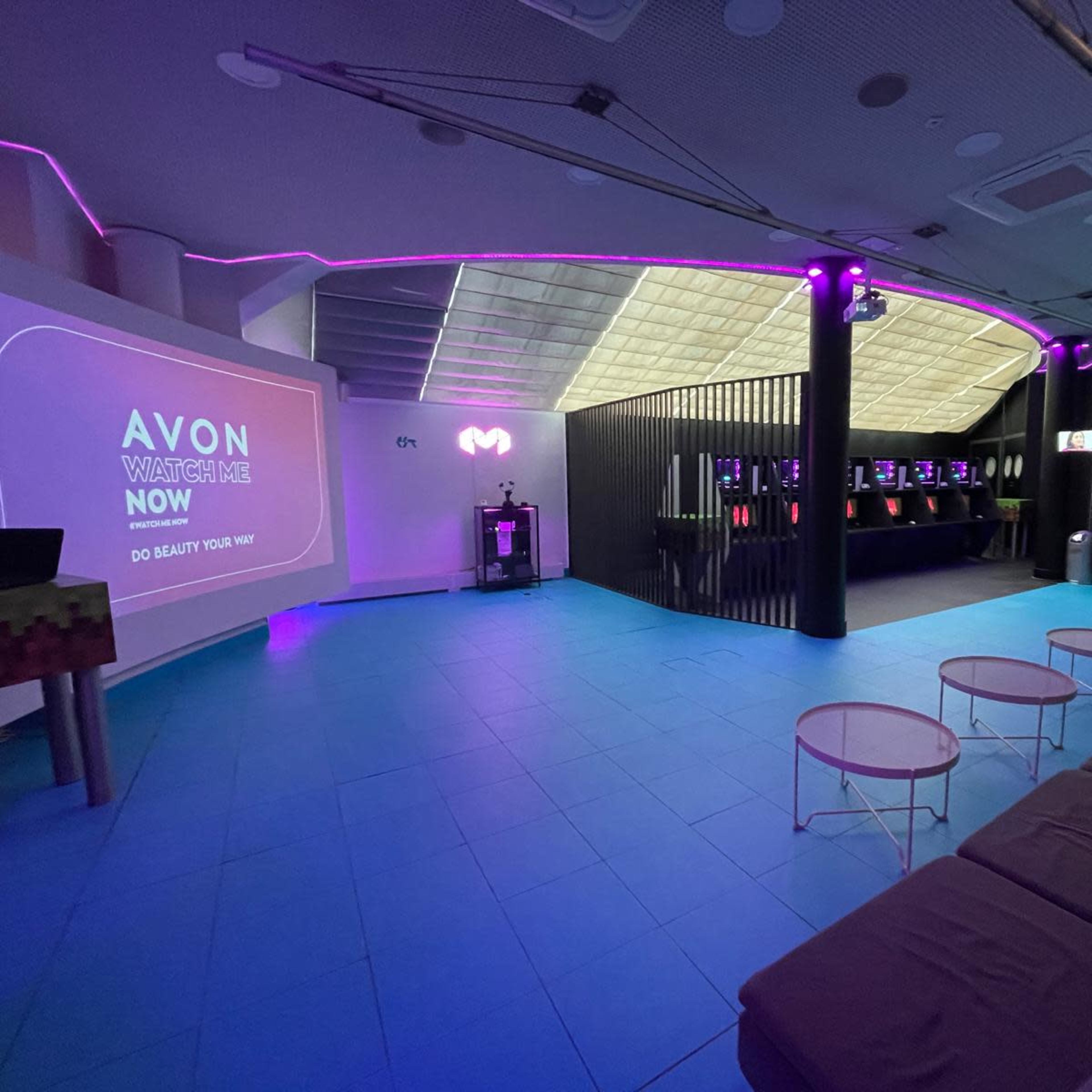 A modern event space with a large presentation screen on one wall, seating area, and a bar-like setup illuminated with colorful lights.