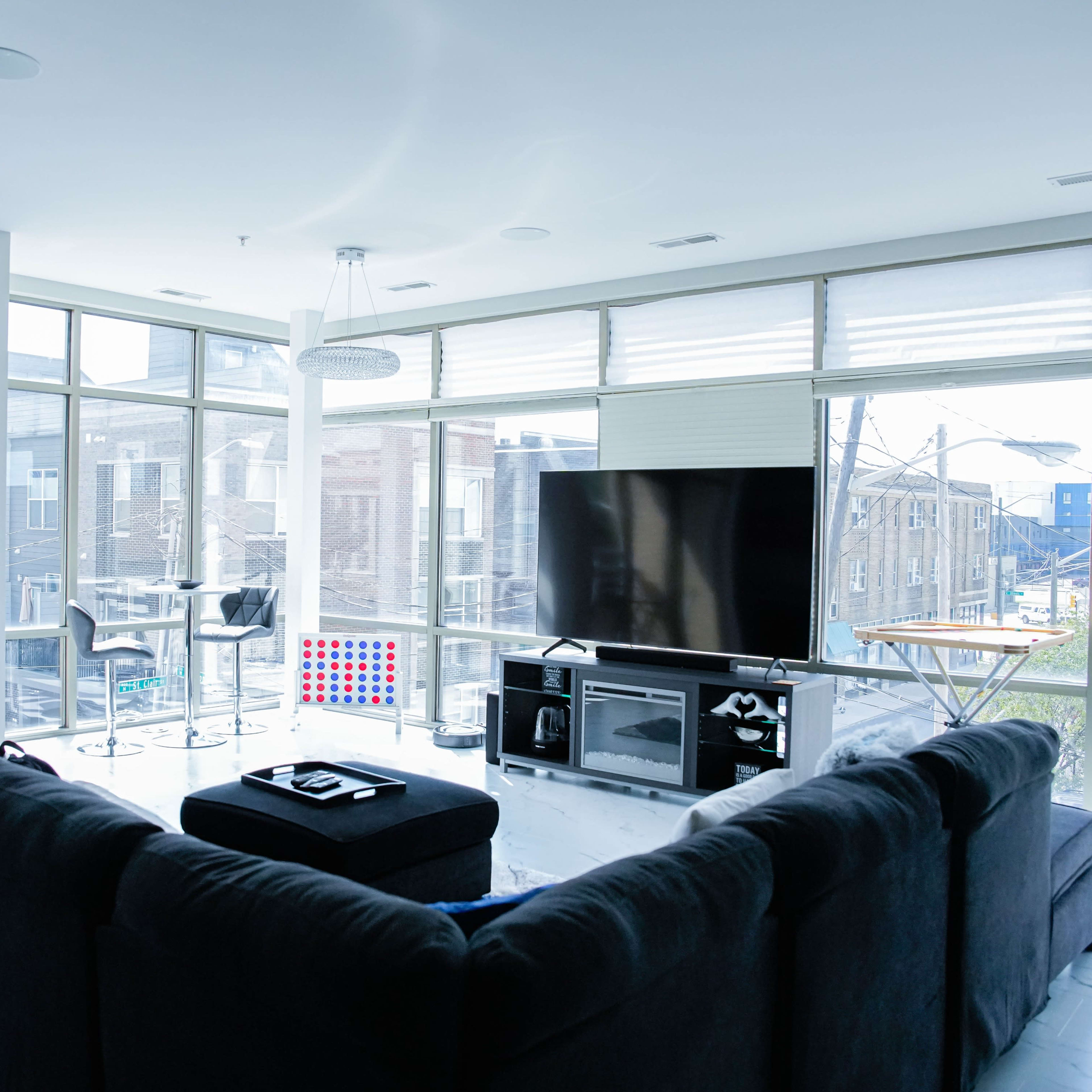 A modern living room with a large sectional sofa facing a flat-screen TV, surrounded by windows that let in natural light.