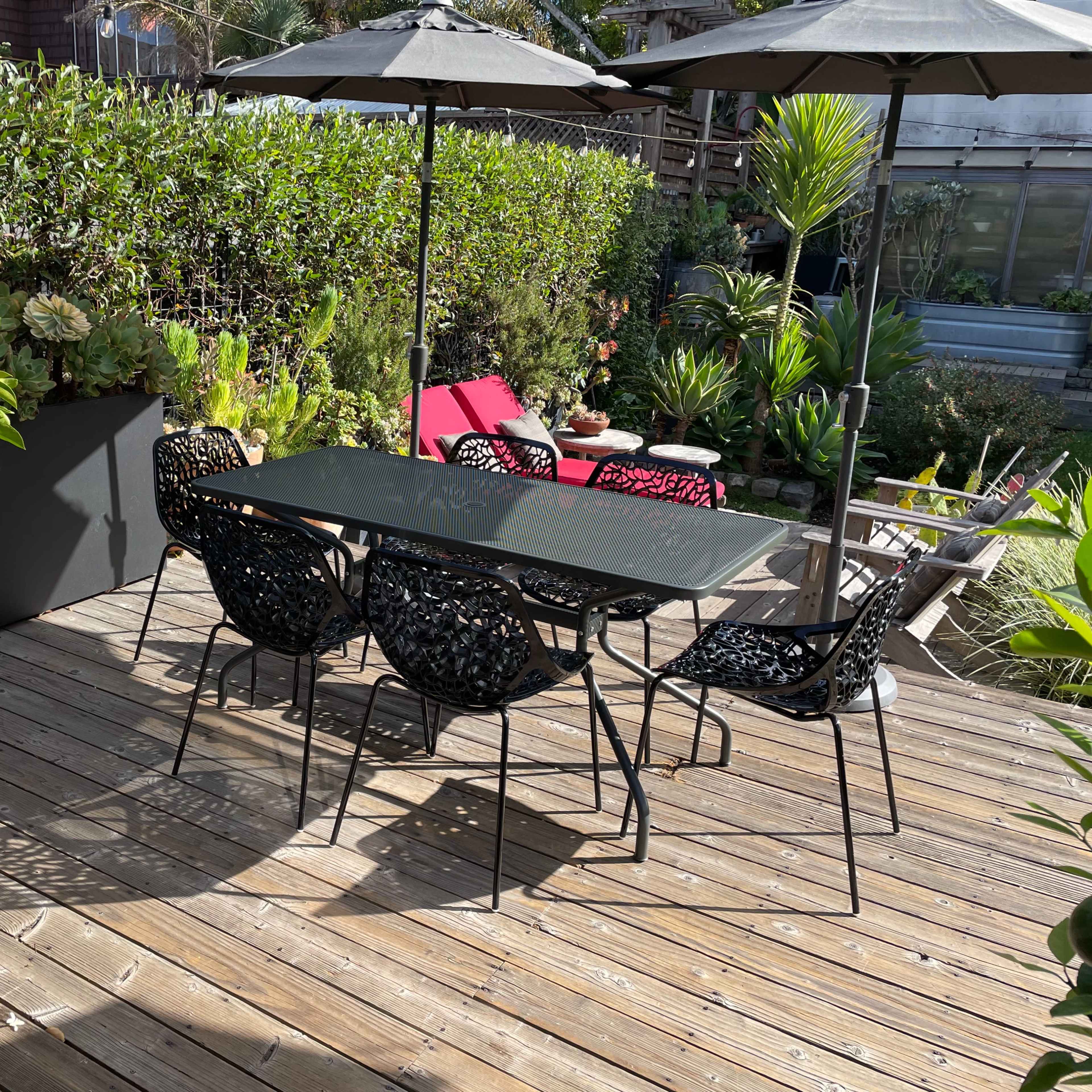 A patio area features a table with four chairs under two umbrellas, surrounded by potted plants and wooden decking.