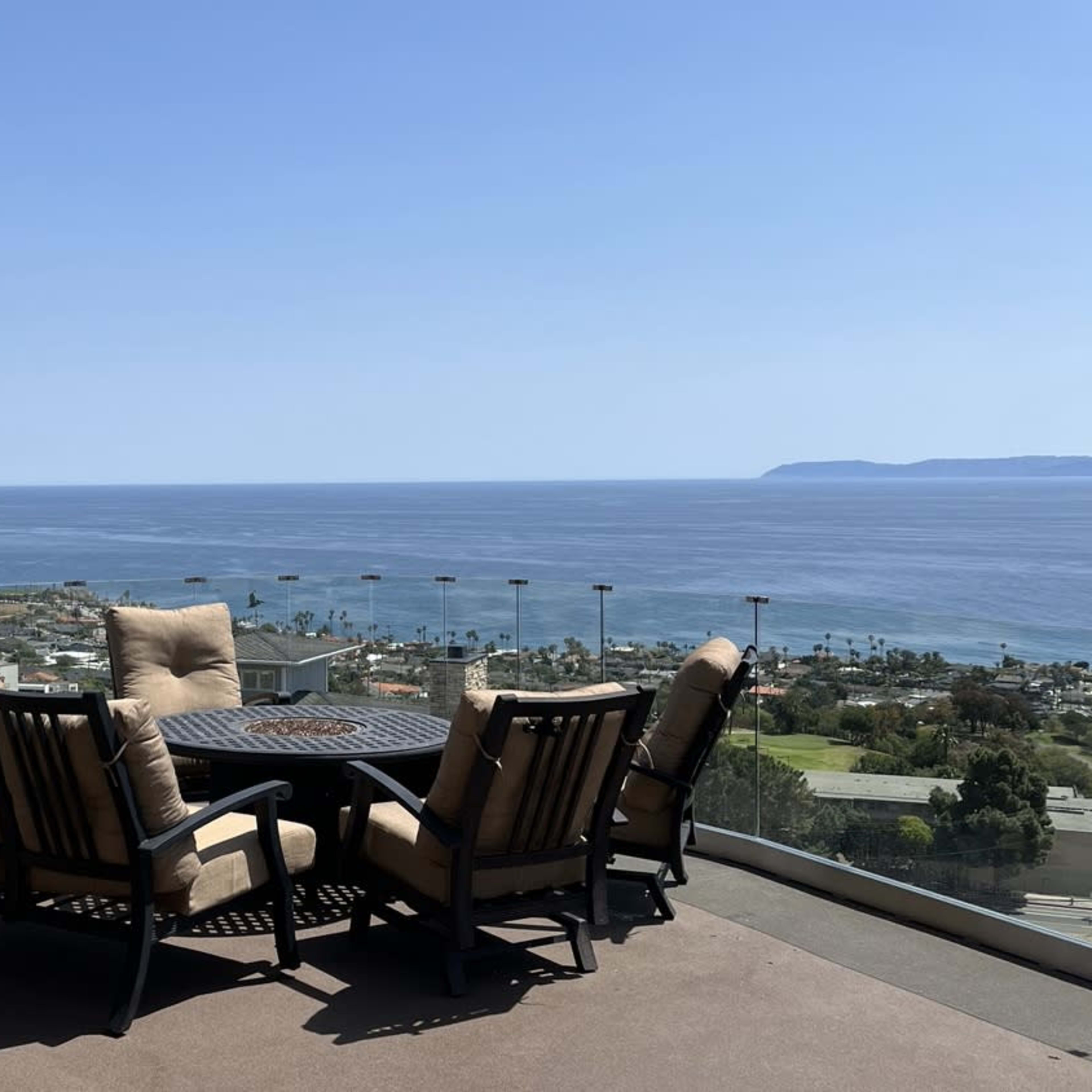 A patio table with four chairs overlooks the ocean and a coastal area under a clear blue sky.