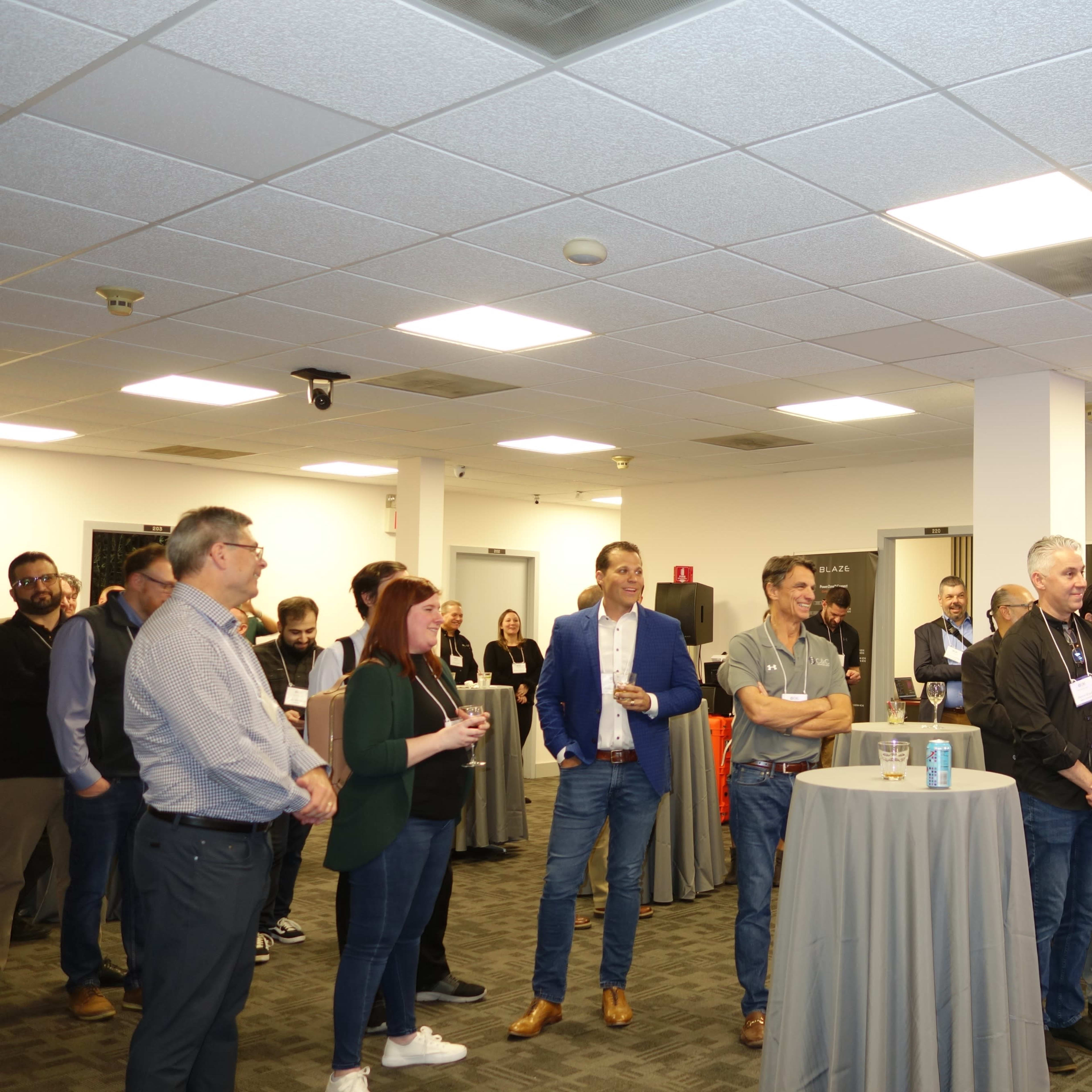 A group of attendees gathers in a conference room, listening to a speaker while holding drinks and standing around a few high tables.
