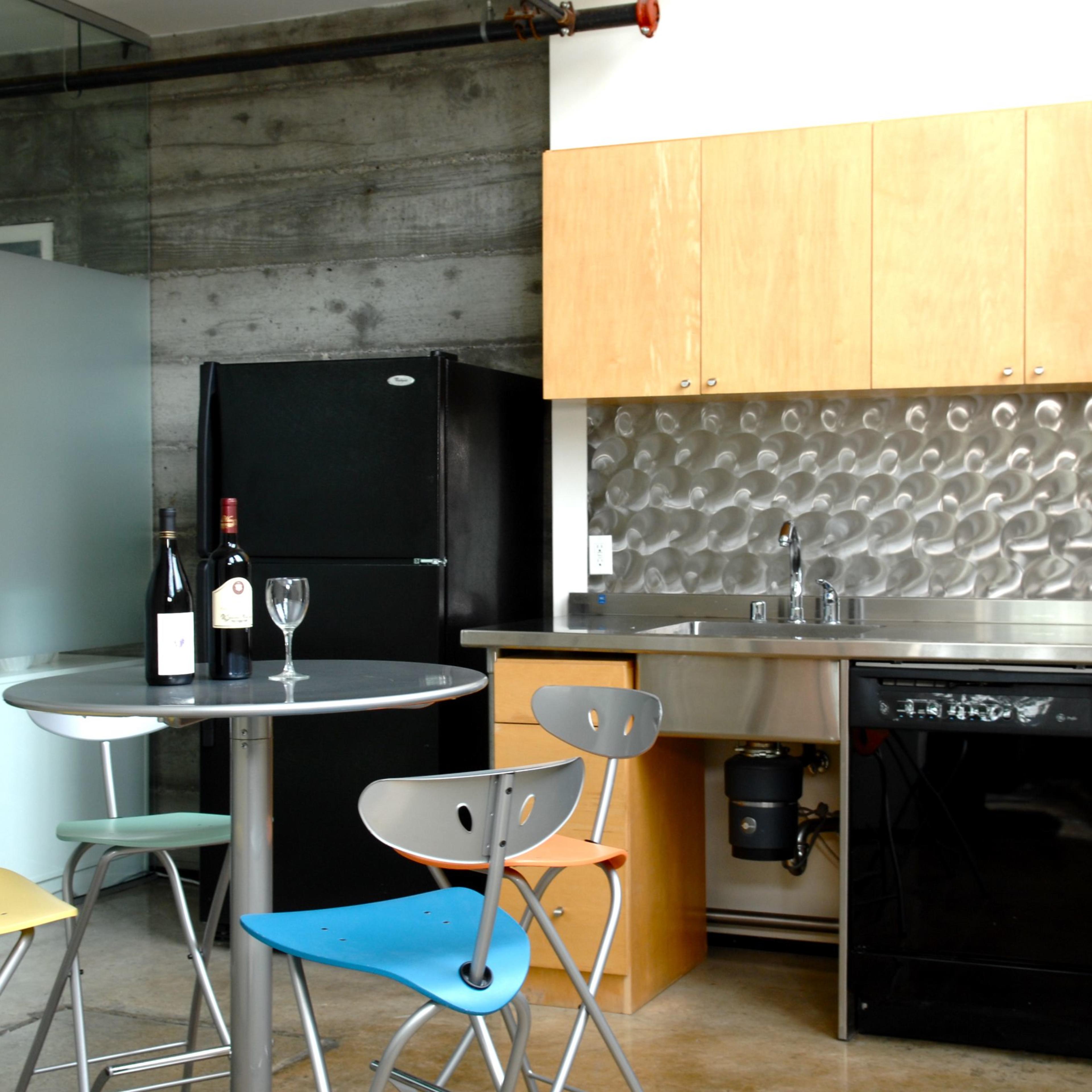 A modern kitchen features stainless steel appliances, wooden cabinetry, and a round dining table with colorful chairs.