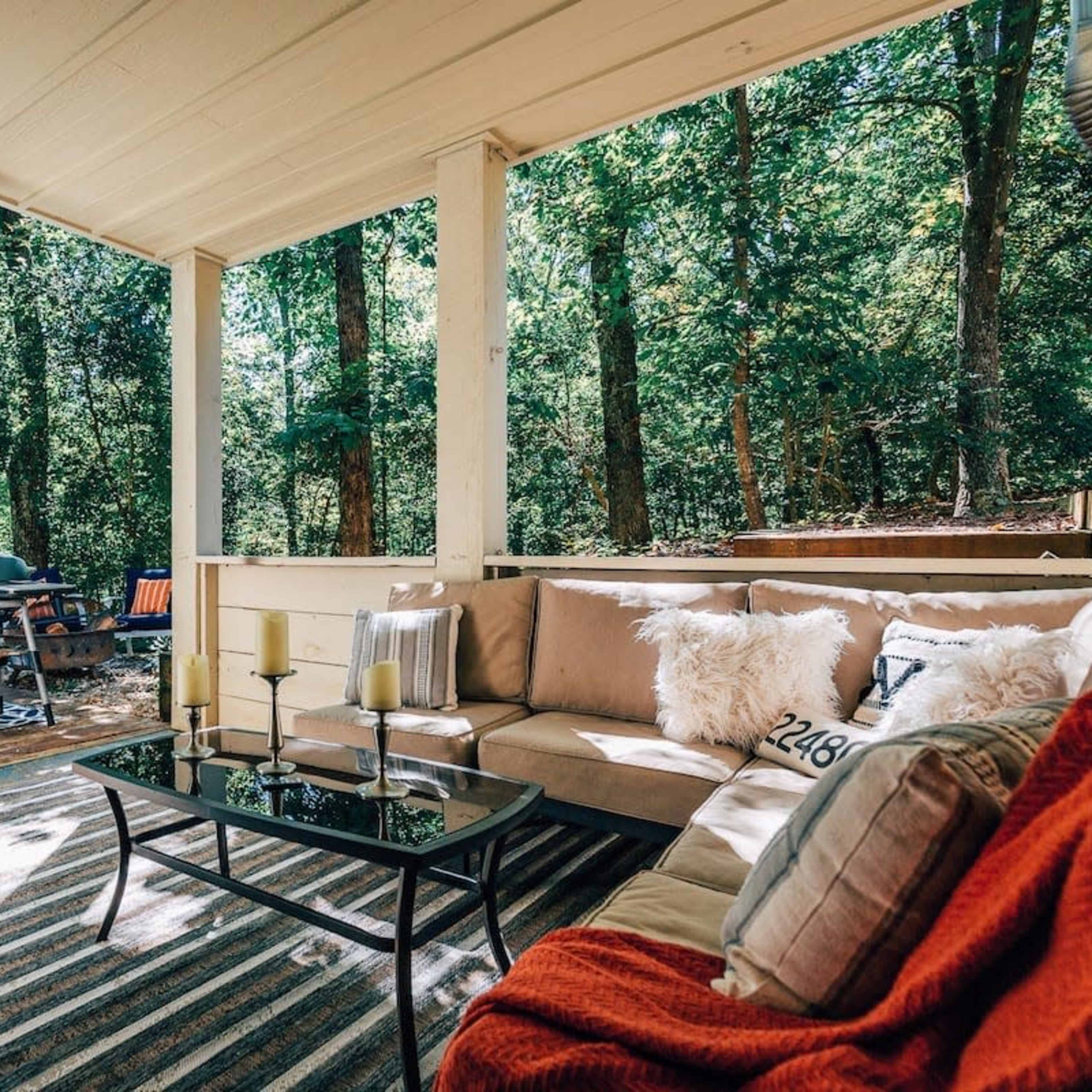 A cozy outdoor seating area features a sectional sofa with cushions, a glass coffee table, and is surrounded by trees with a hot tub visible nearby.