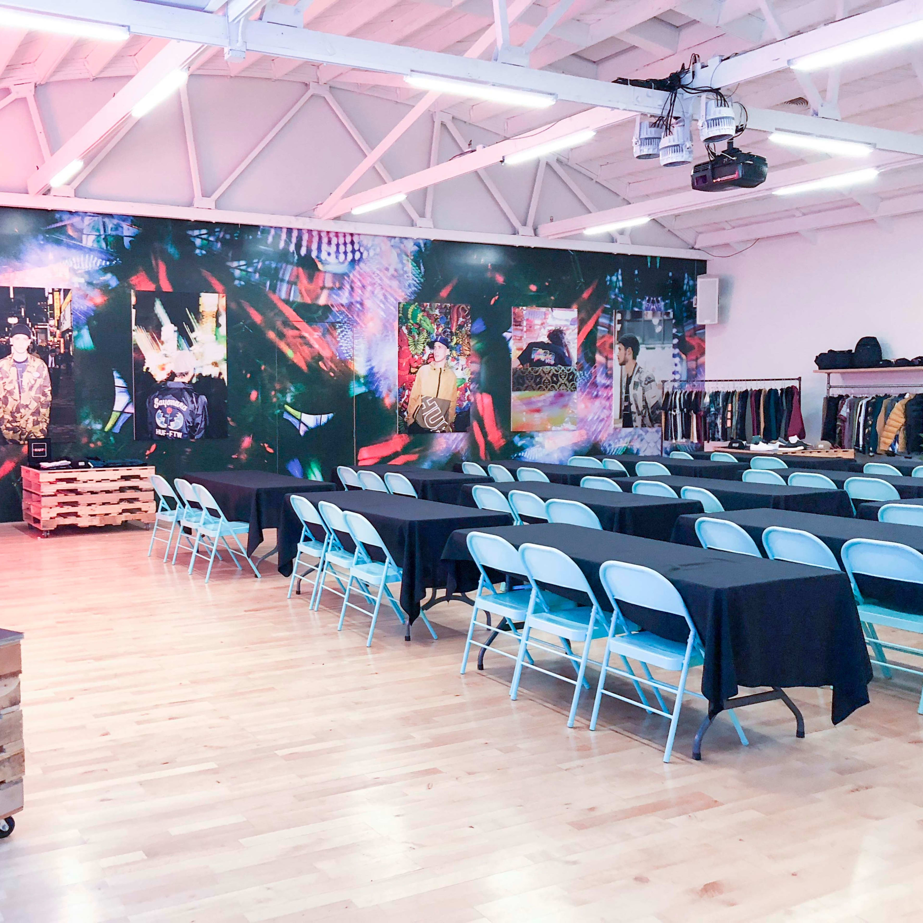 A spacious room features multiple tables covered with black cloth, surrounded by blue chairs, against a vividly decorated wall adorned with colorful graphics and images.