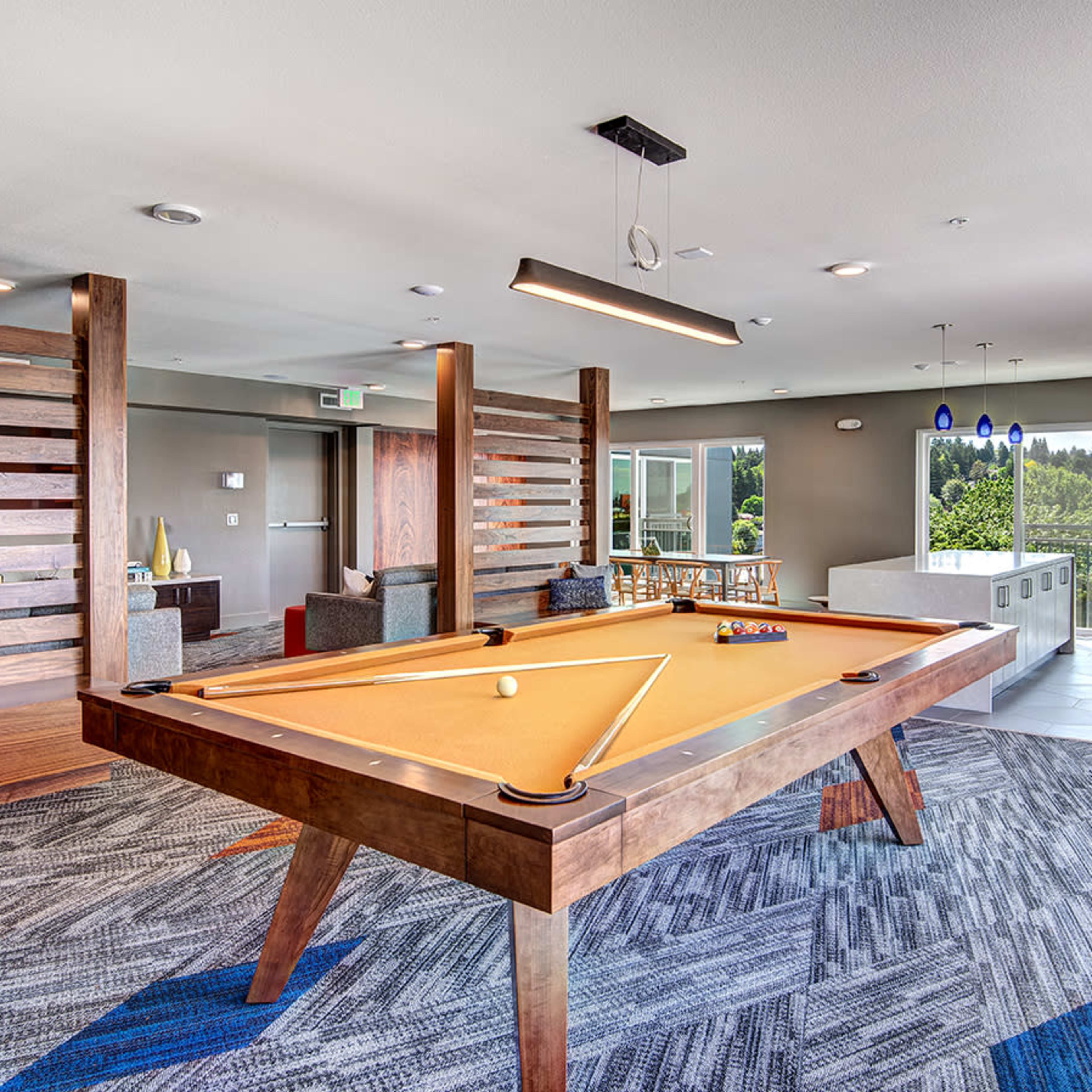 A modern lounge area features a wooden pool table surrounded by seating and large windows that provide natural light and a view of the outdoors.