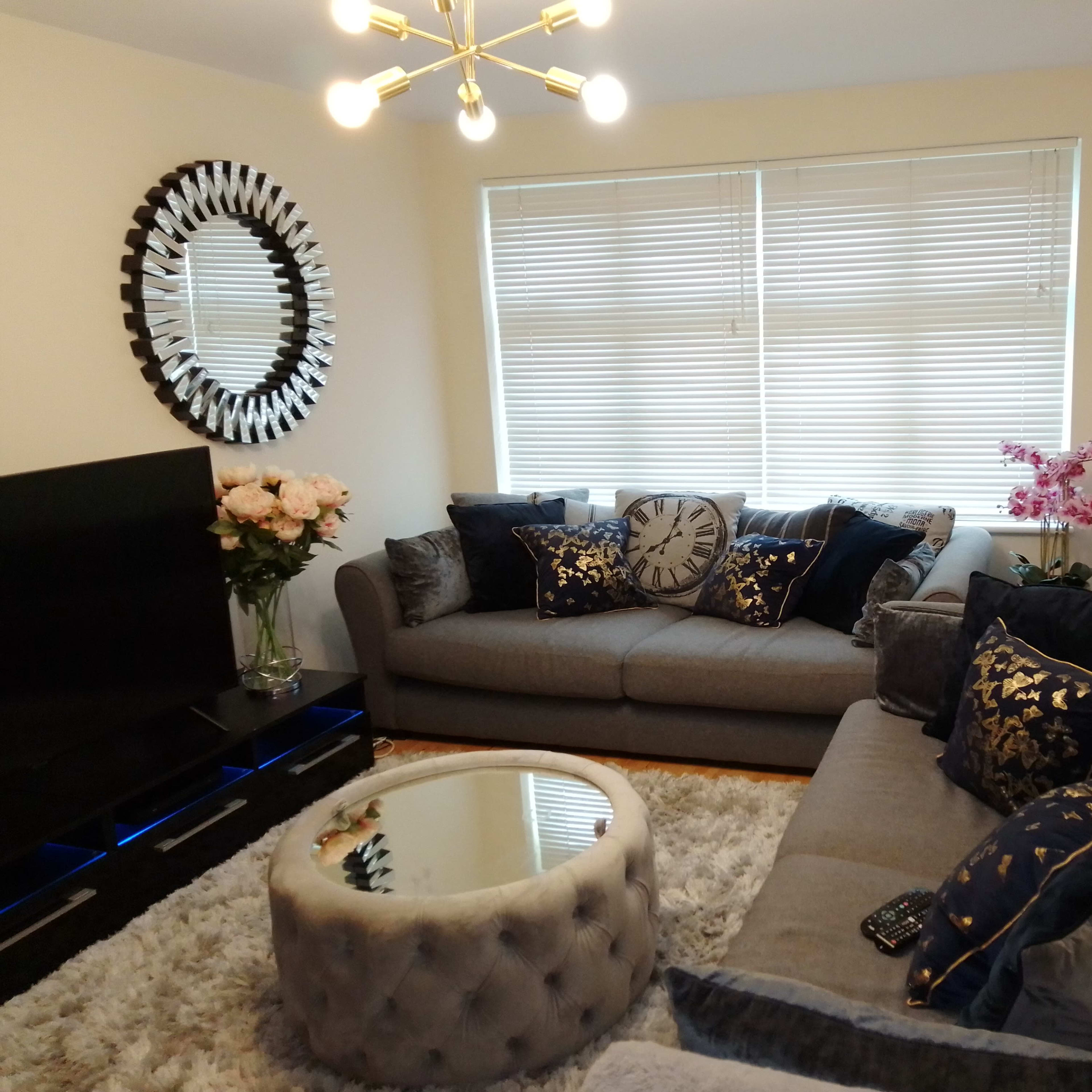 A modern living room featuring a gray sectional sofa, a round coffee table, a large mirror on the wall, and decorative pillows, with a floral arrangement nearby.