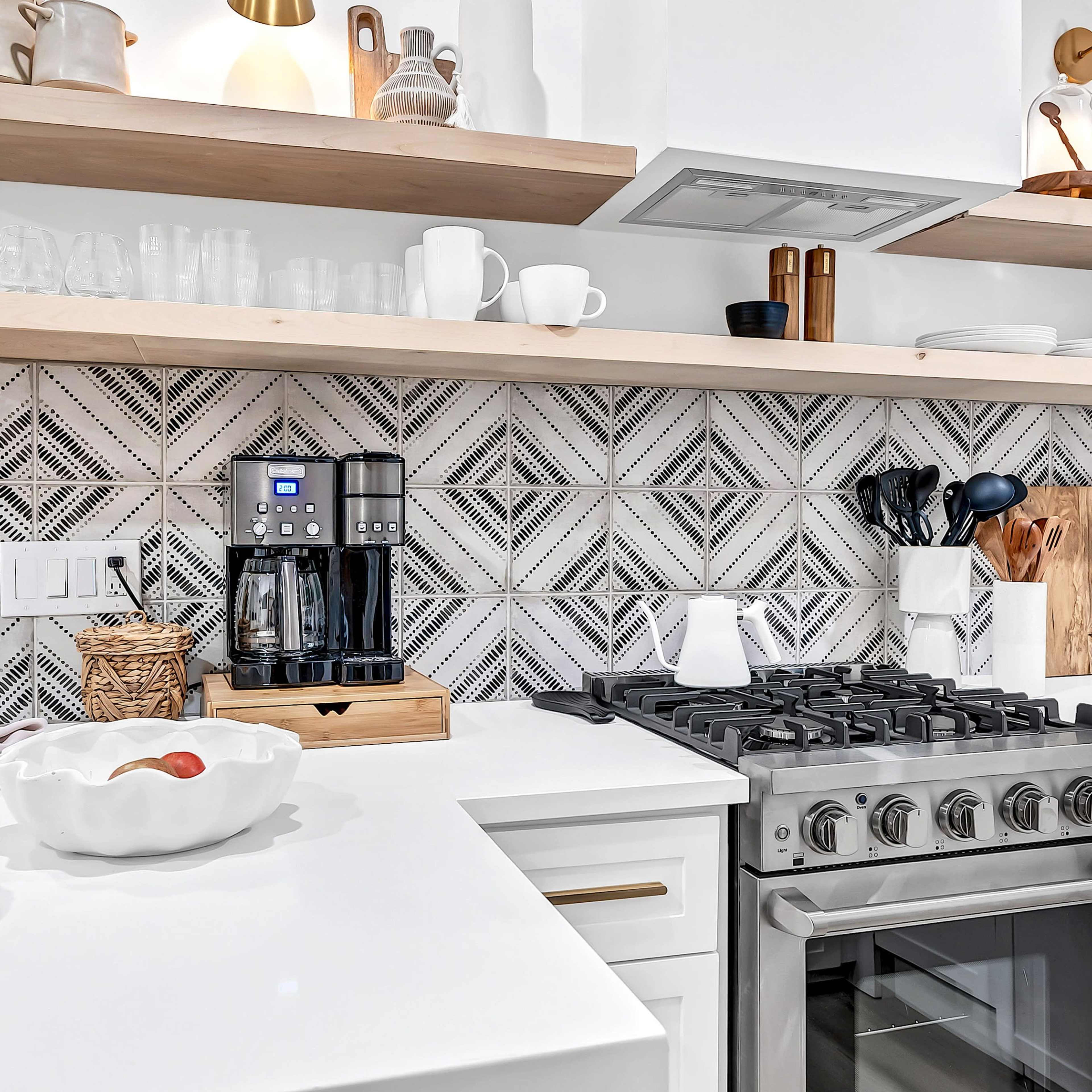 A modern kitchen featuring a sleek gas stove, a coffee maker, and open shelves displaying dishware and decorative items against a patterned tile backsplash.