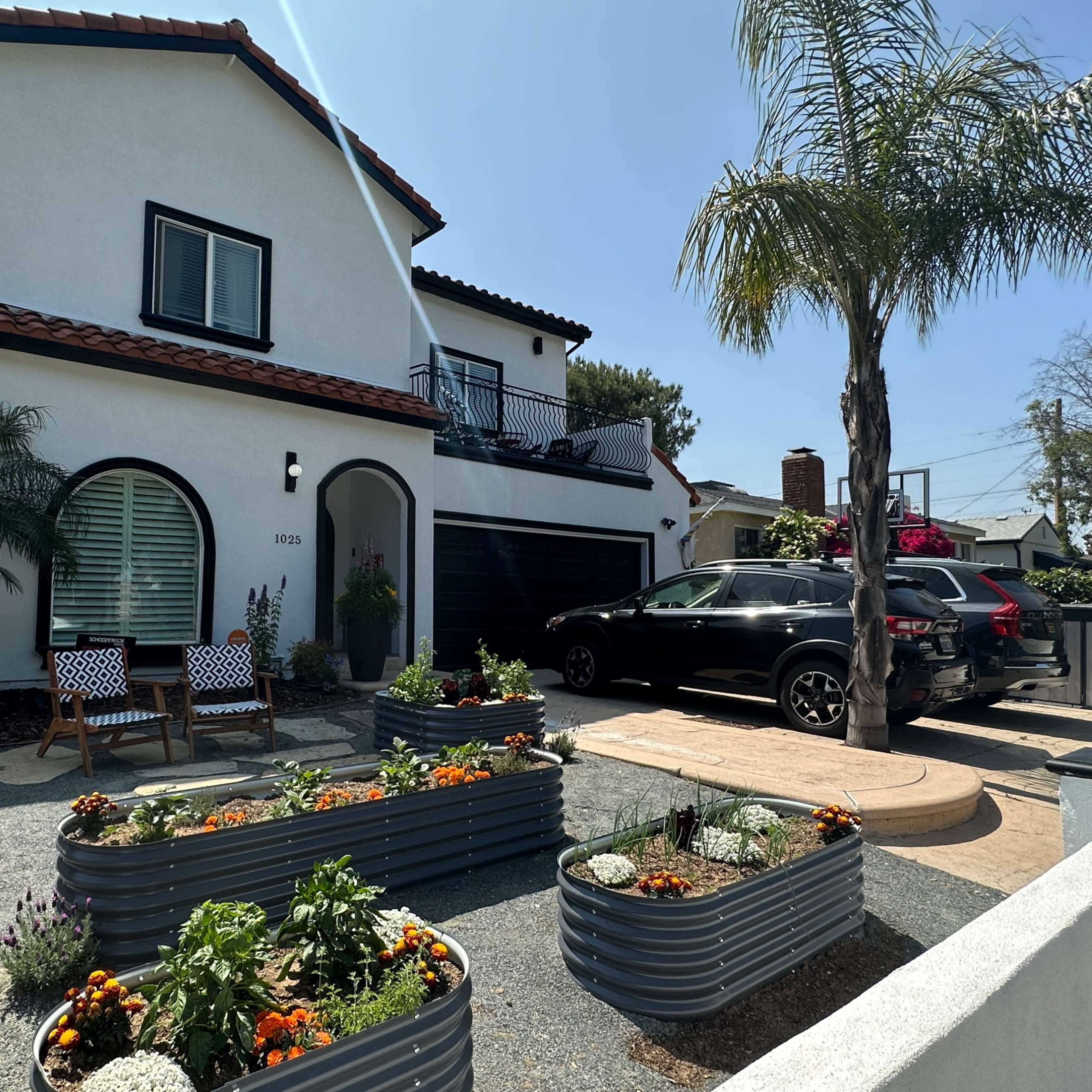 A two-story white house with a tiled roof is surrounded by landscaped gardens featuring raised metal planters and a black vehicle parked in the driveway.