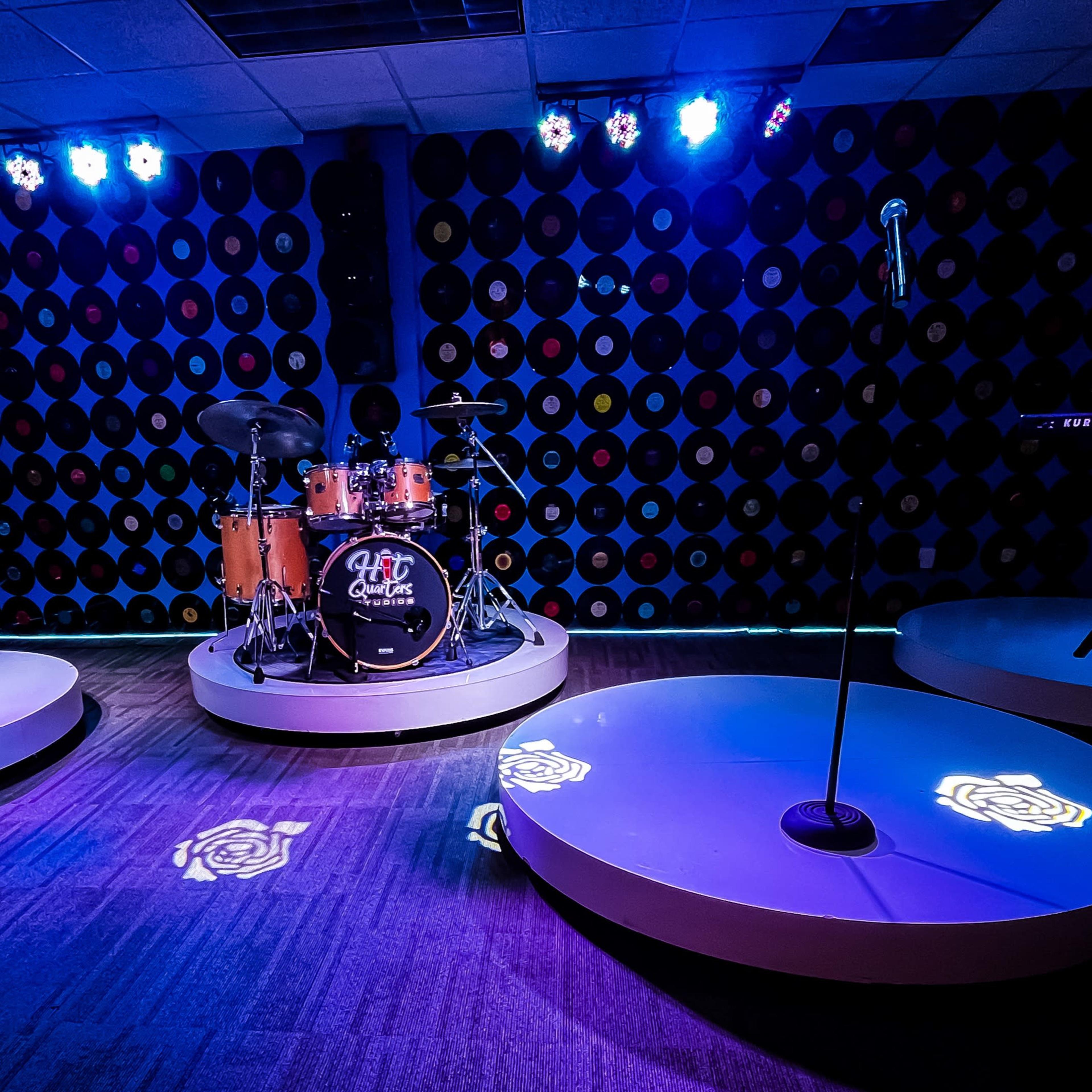 A music rehearsal space featuring a drum set, keyboard, and microphone stands arranged on circular platforms, illuminated by blue stage lighting against a backdrop of vinyl records.