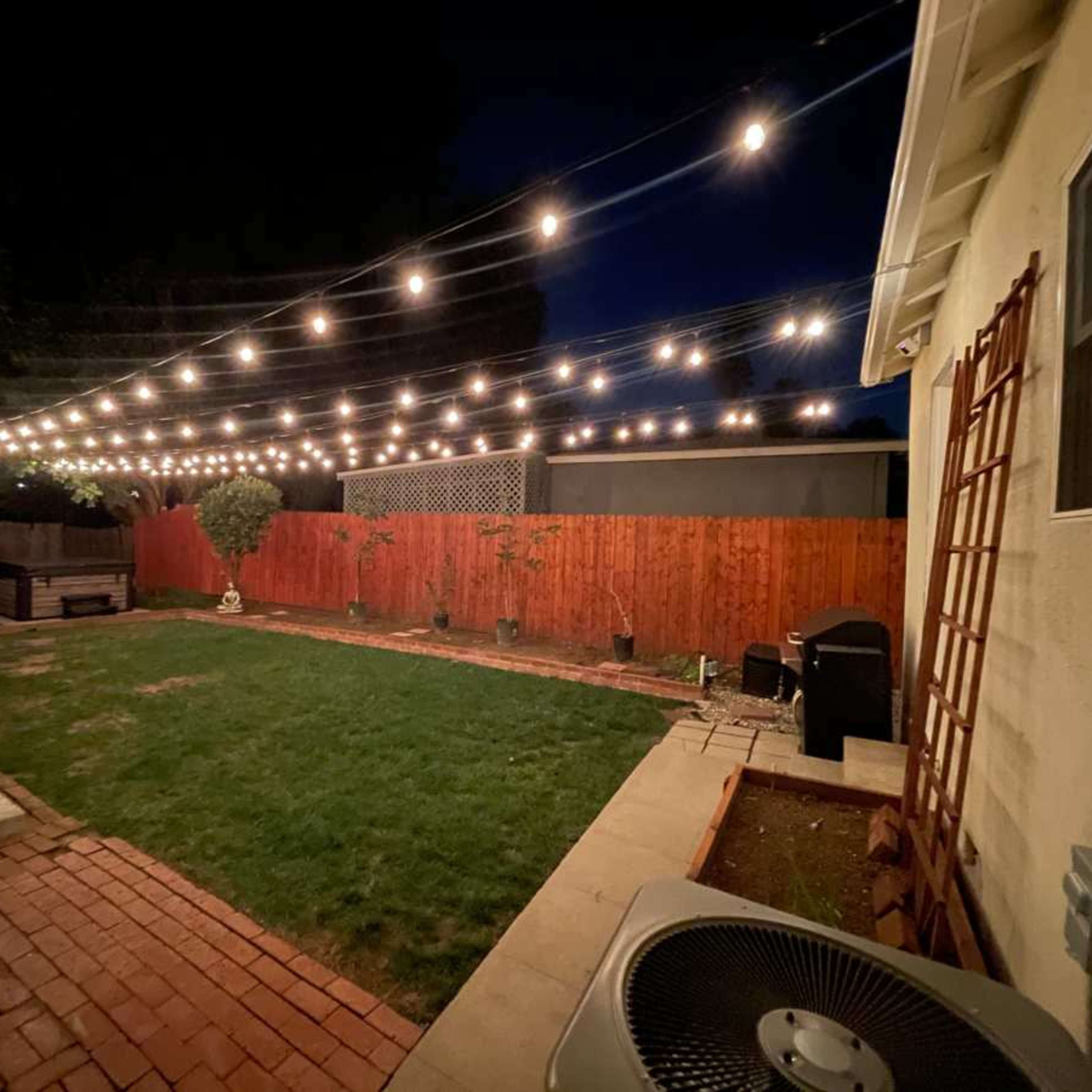 A backyard at night features string lights overhead, a grassy area, brick and concrete paths, and wooden fencing.