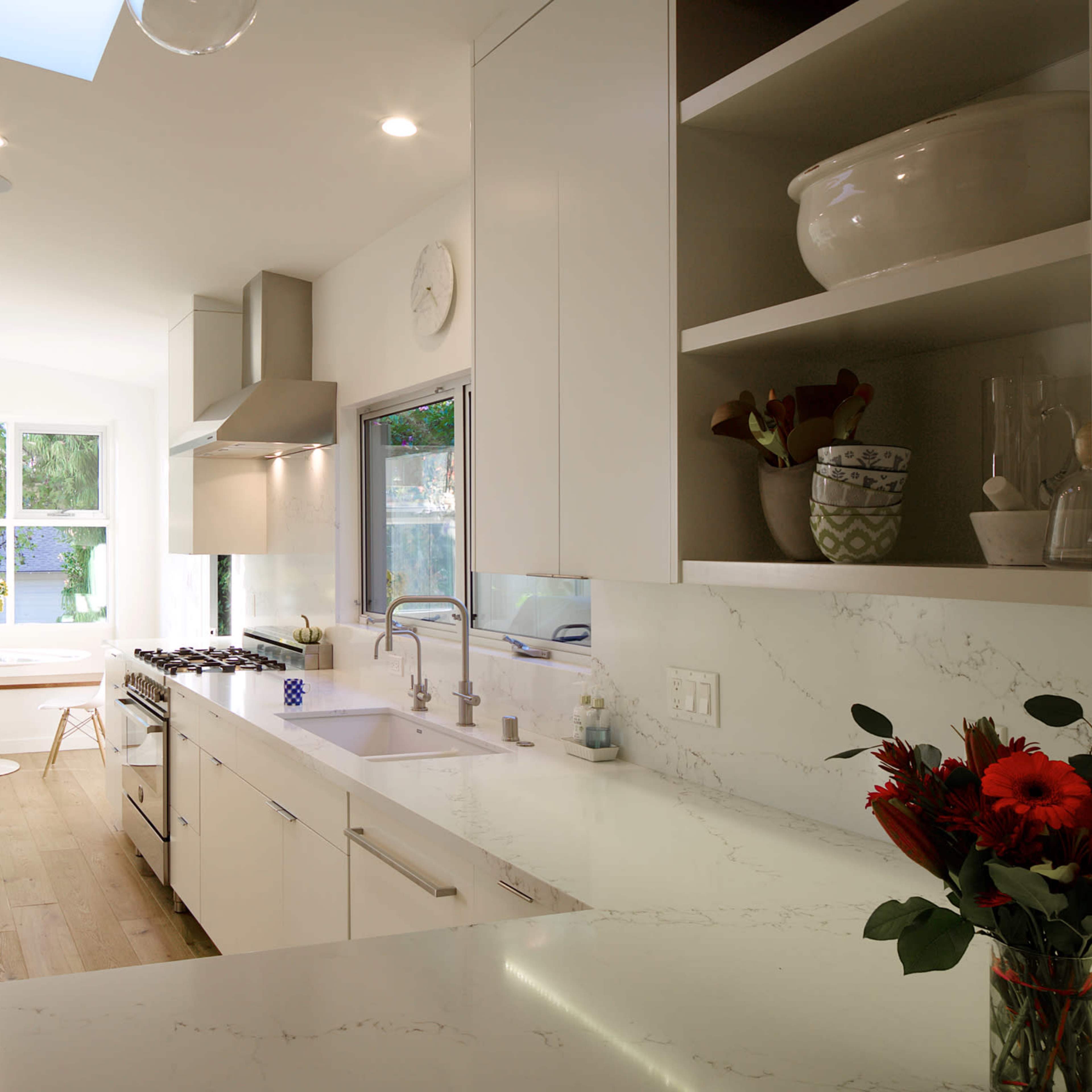 A sleek, modern kitchen features white cabinetry, a marble countertop, and a view of a sunlit dining area through large windows.
