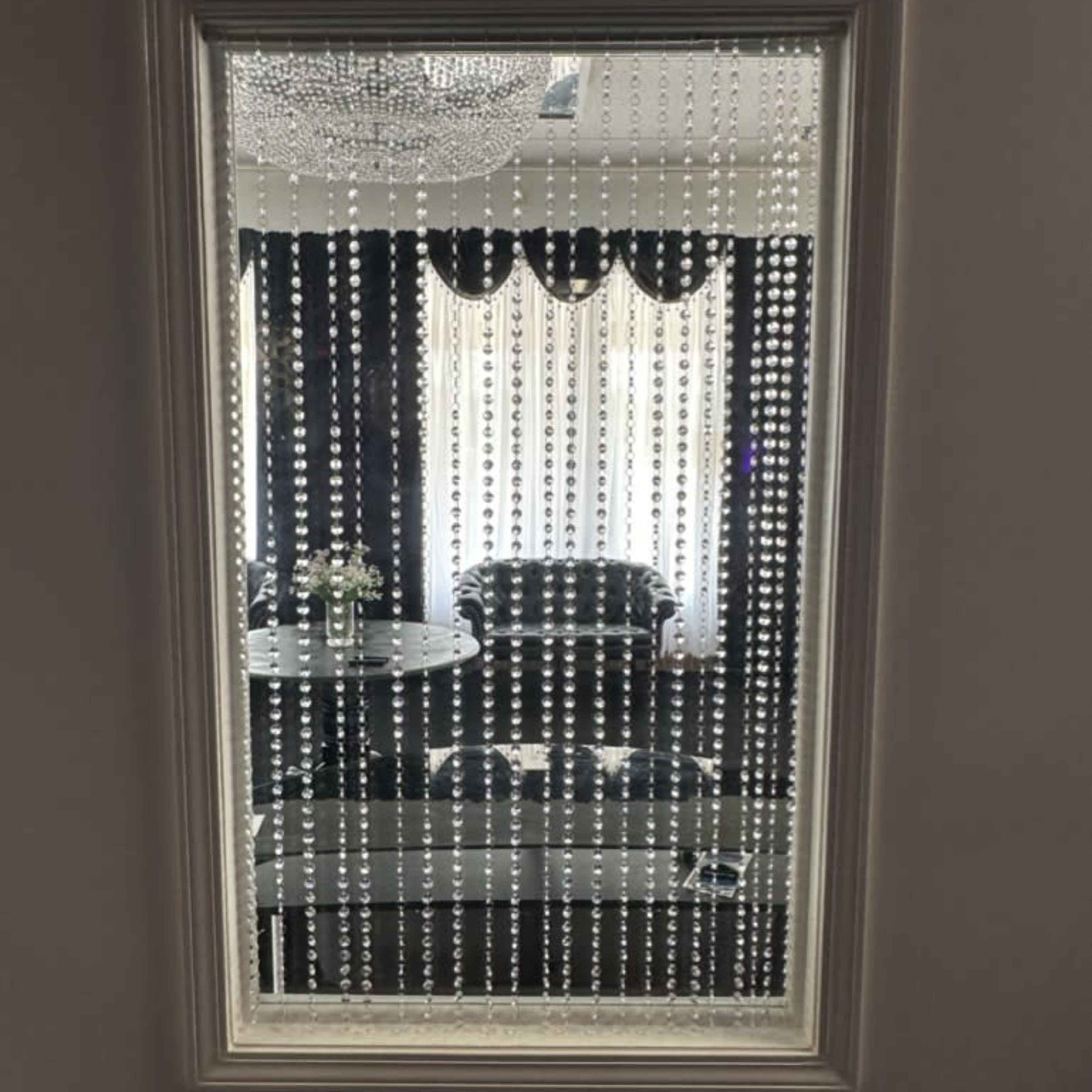 A framed mirror reflects a room with a chandelier, a dark sofa, and a table, all partially obscured by hanging beads.