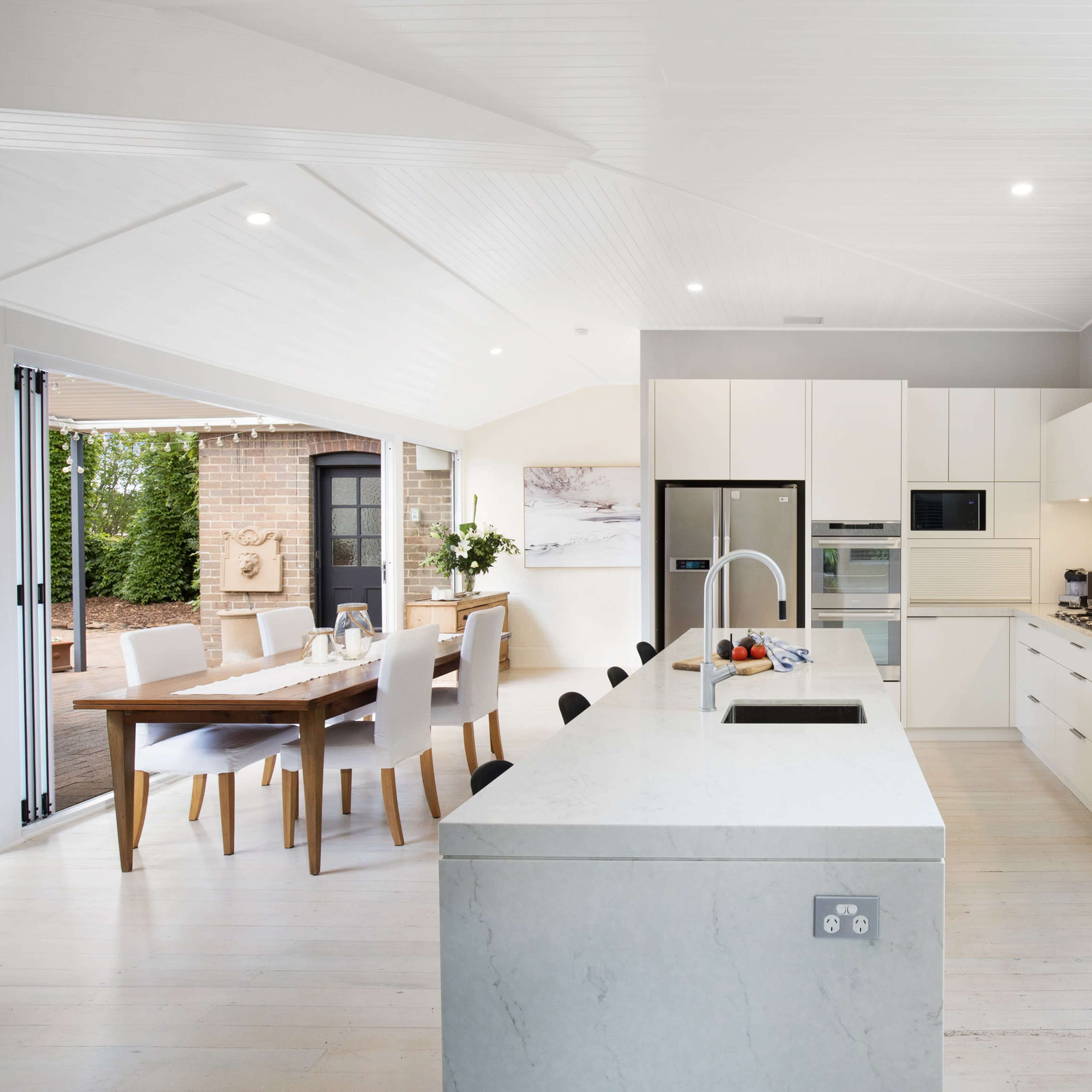 A modern kitchen featuring white cabinetry, stainless steel appliances, and an island with a double sink, adjacent to a dining area with a wooden table.
