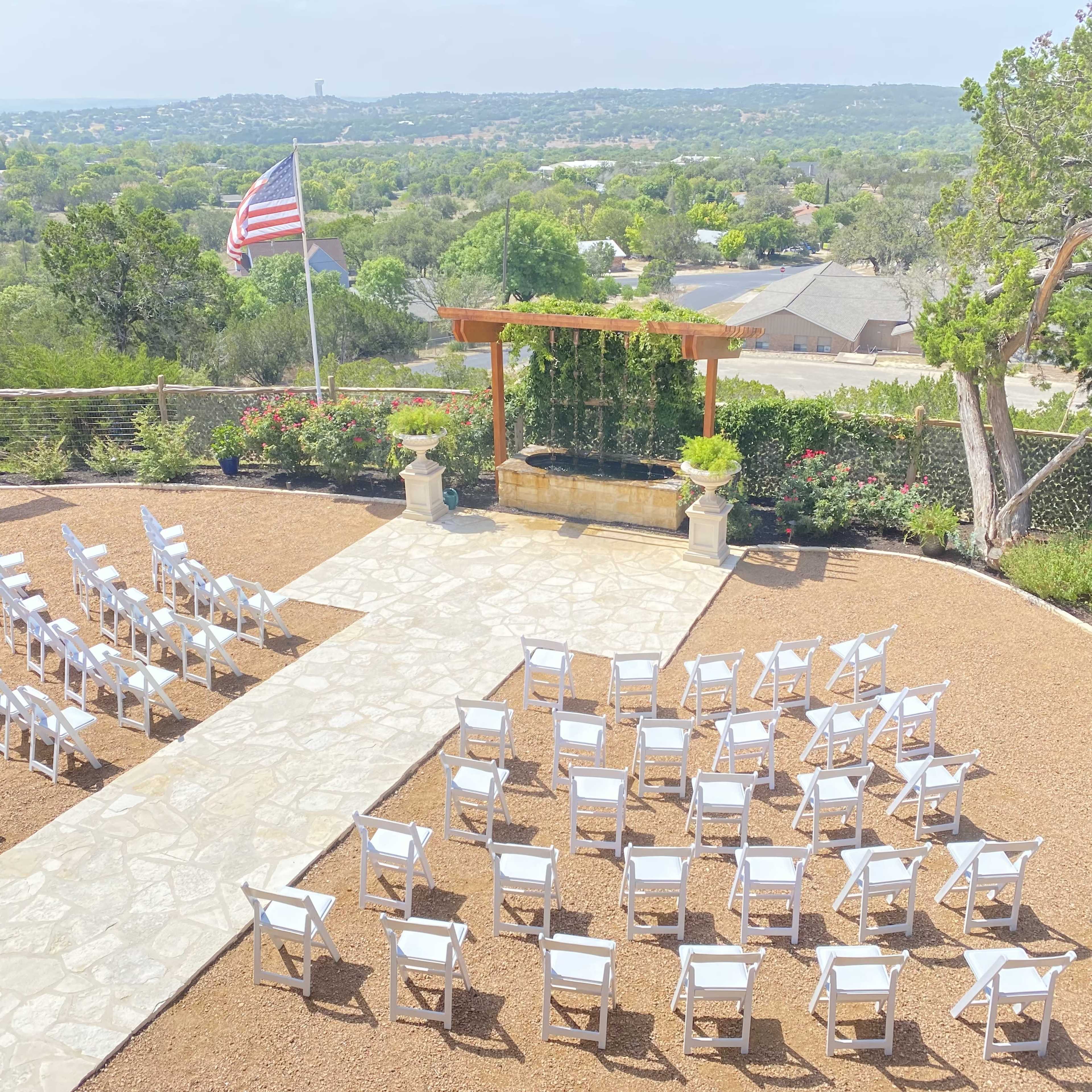 A row of white chairs is arranged on a stone pathway for an outdoor event, overlooking a green landscape with a flagpole in the background.