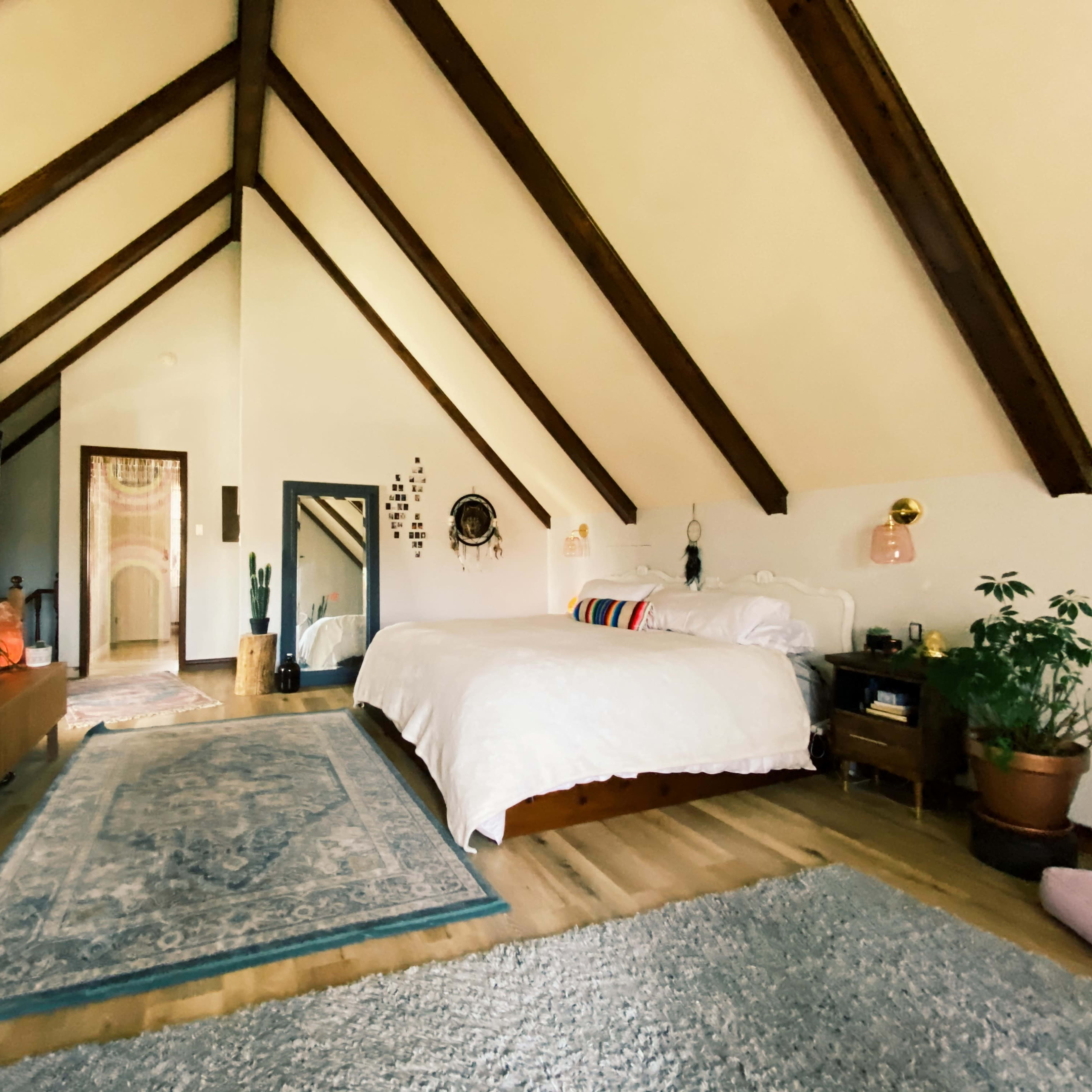 A spacious bedroom with a high, vaulted ceiling, featuring a large bed, a television mounted on the wall, and plants placed around the room.