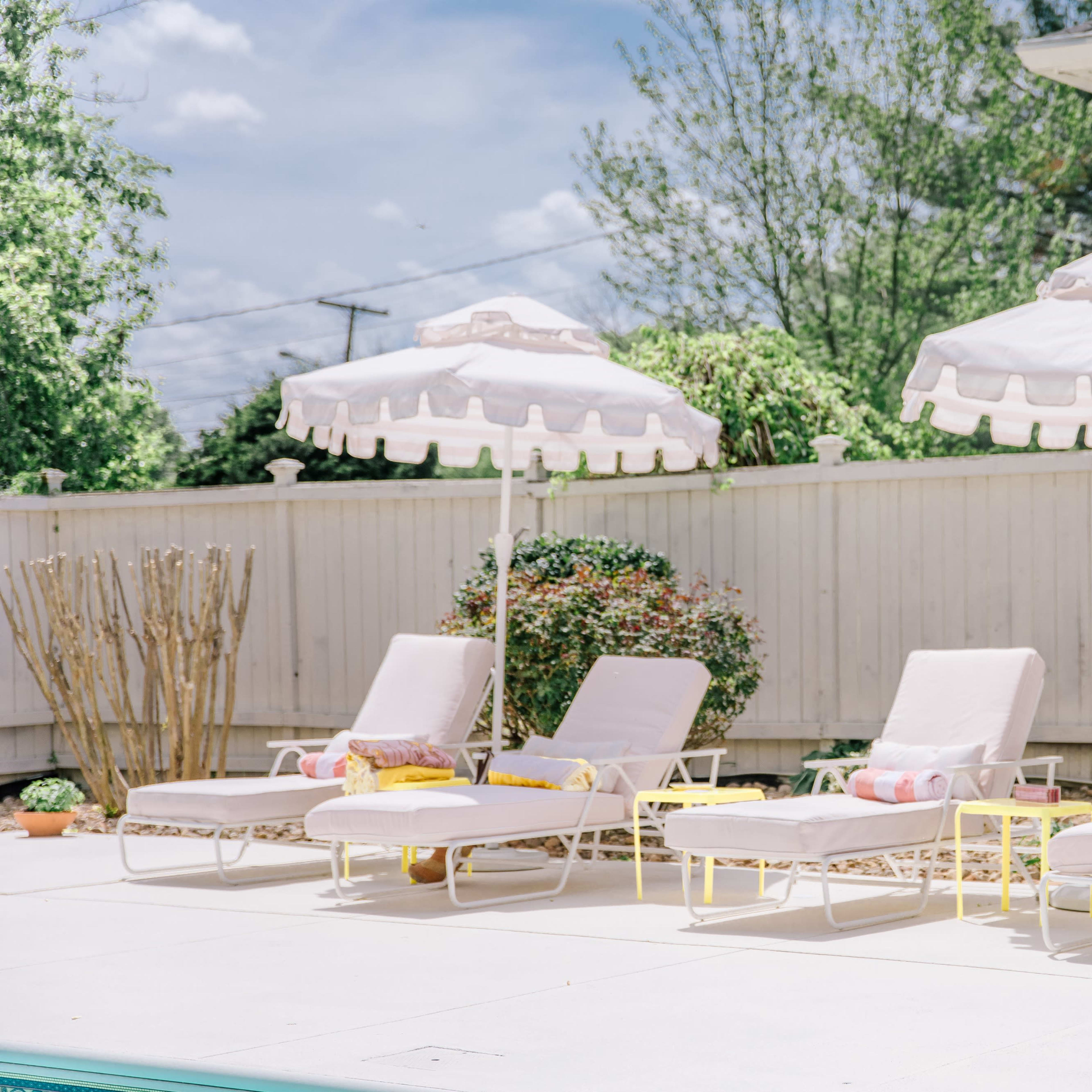 A row of four white lounge chairs with pink cushions and matching umbrellas beside a pool, set against a landscaped backdrop.