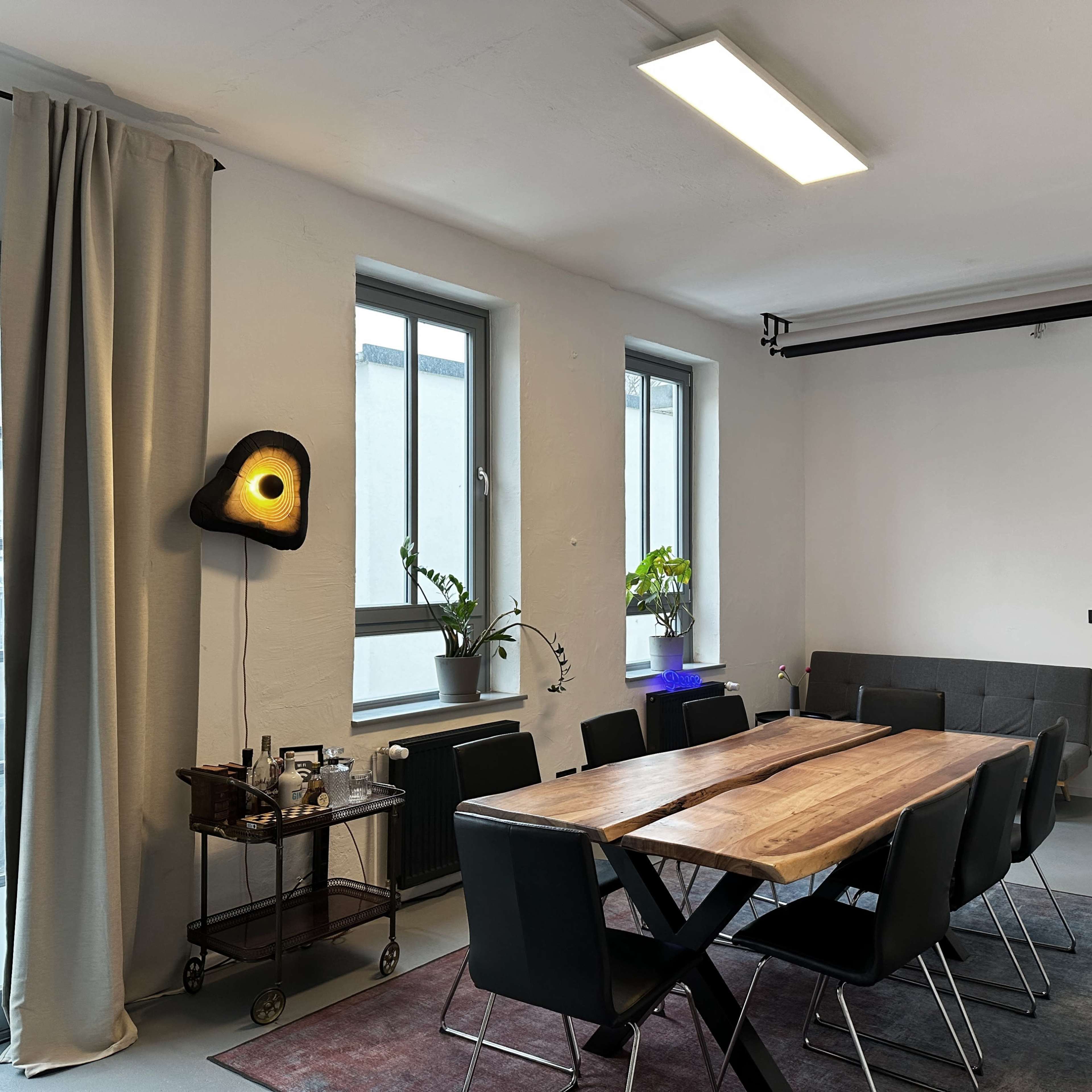 A spacious room features a long wooden dining table surrounded by black chairs, with large windows allowing natural light and a cart displaying beverages in the corner.