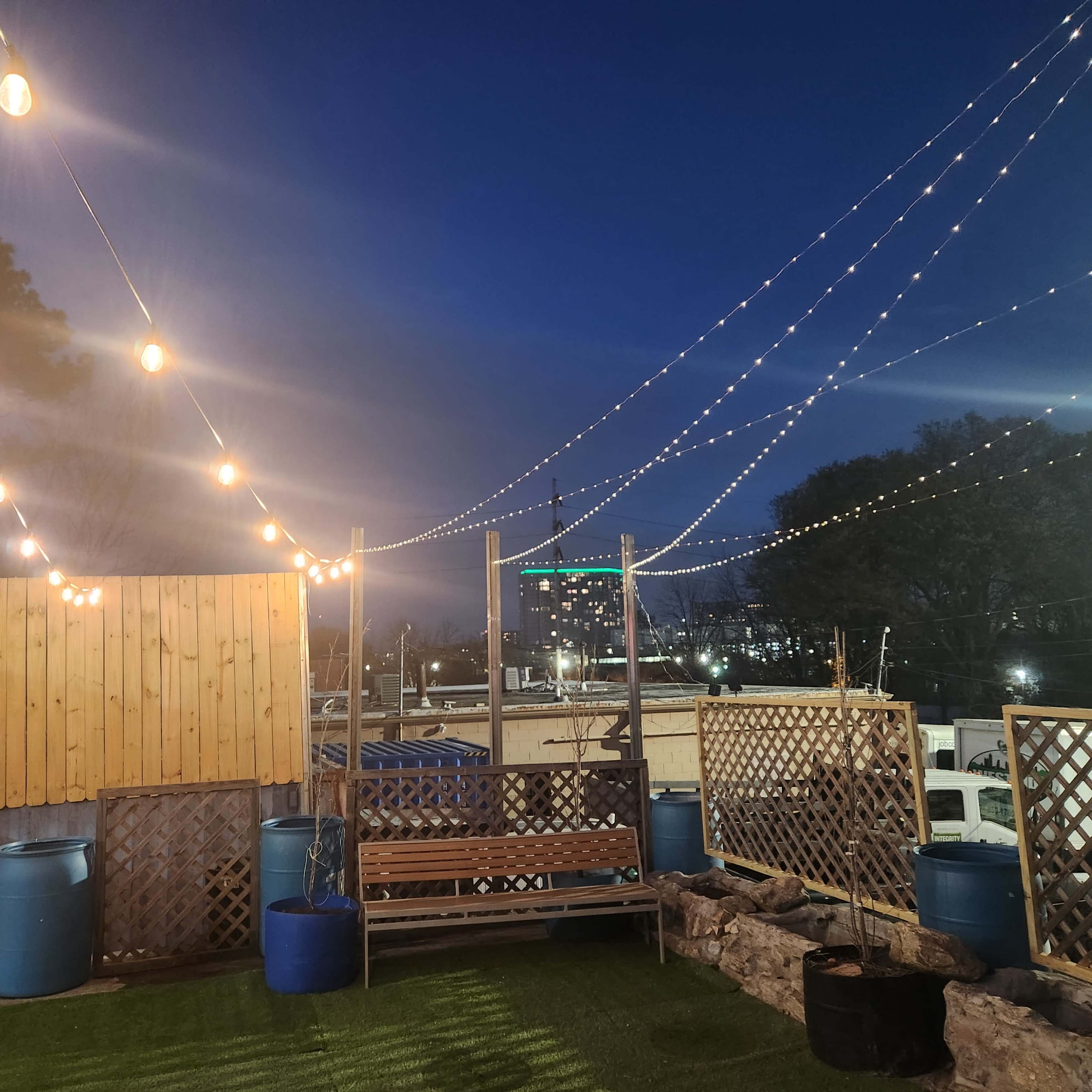 An outdoor area with string lights overhead, wooden fences, and seating, set against a twilight sky.