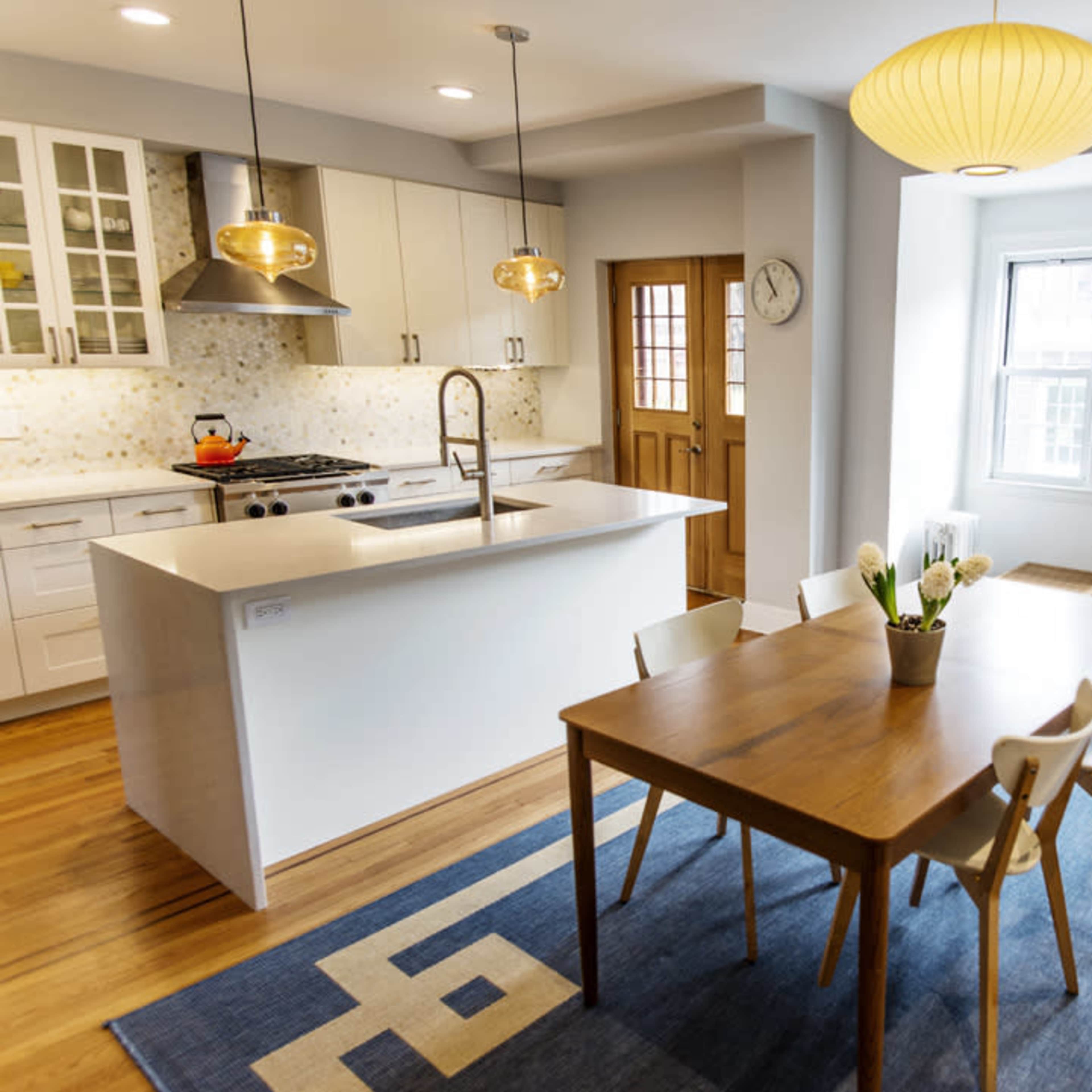 A modern kitchen and dining area features a wooden table set with chairs, pendant lighting, and large windows allowing natural light to fill the space.