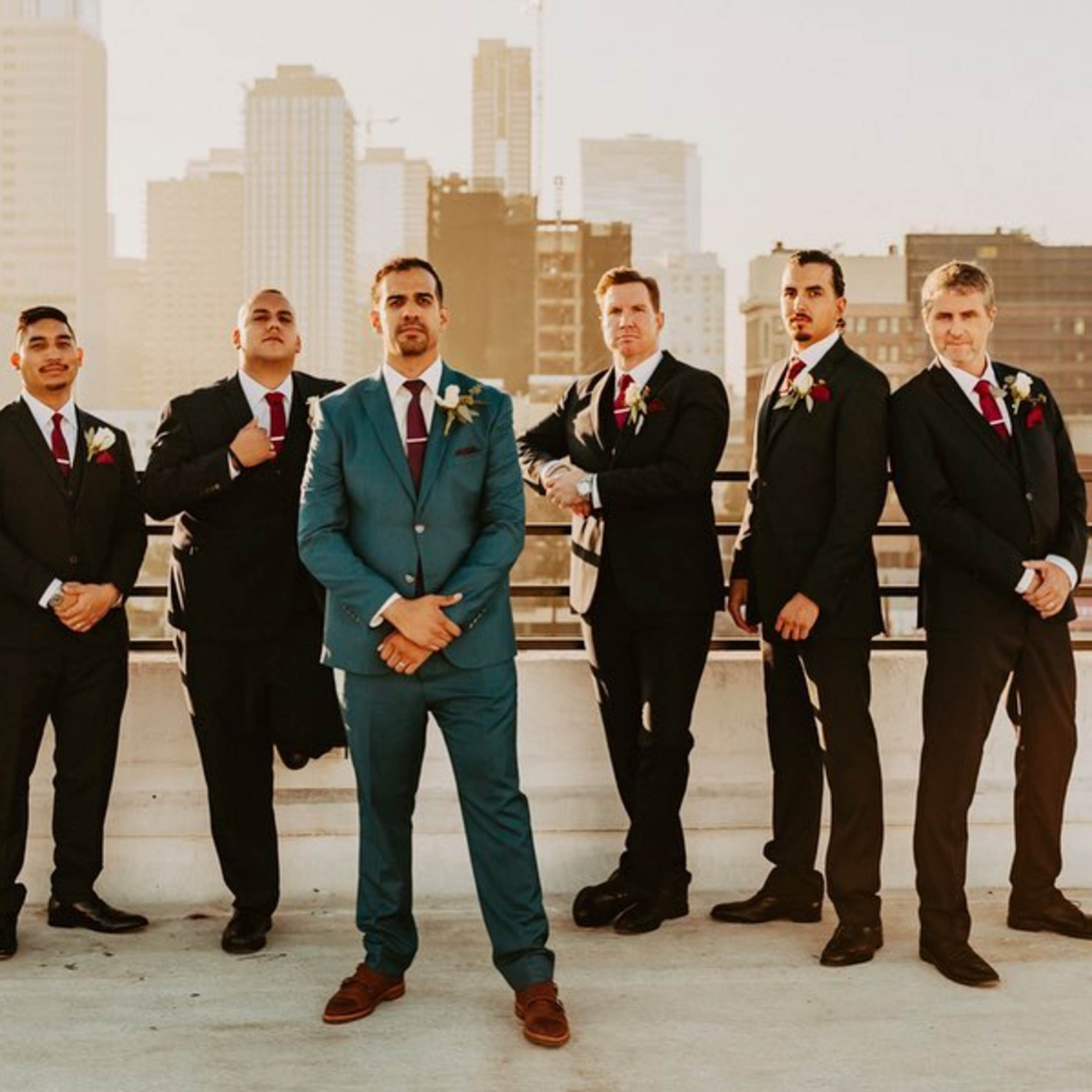 A group of seven men dressed in suits stands on a rooftop with a city skyline in the background.