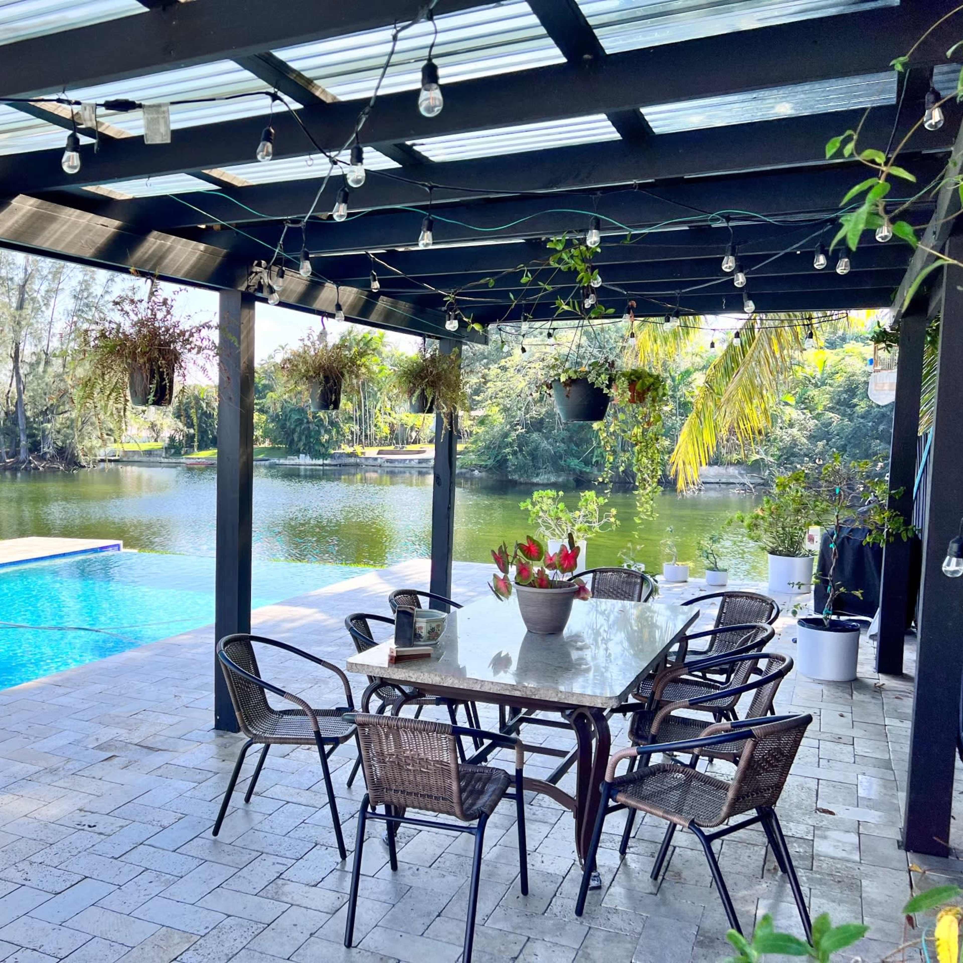 A covered patio with a dining table and chairs overlooks a swimming pool and a calm body of water surrounded by greenery.