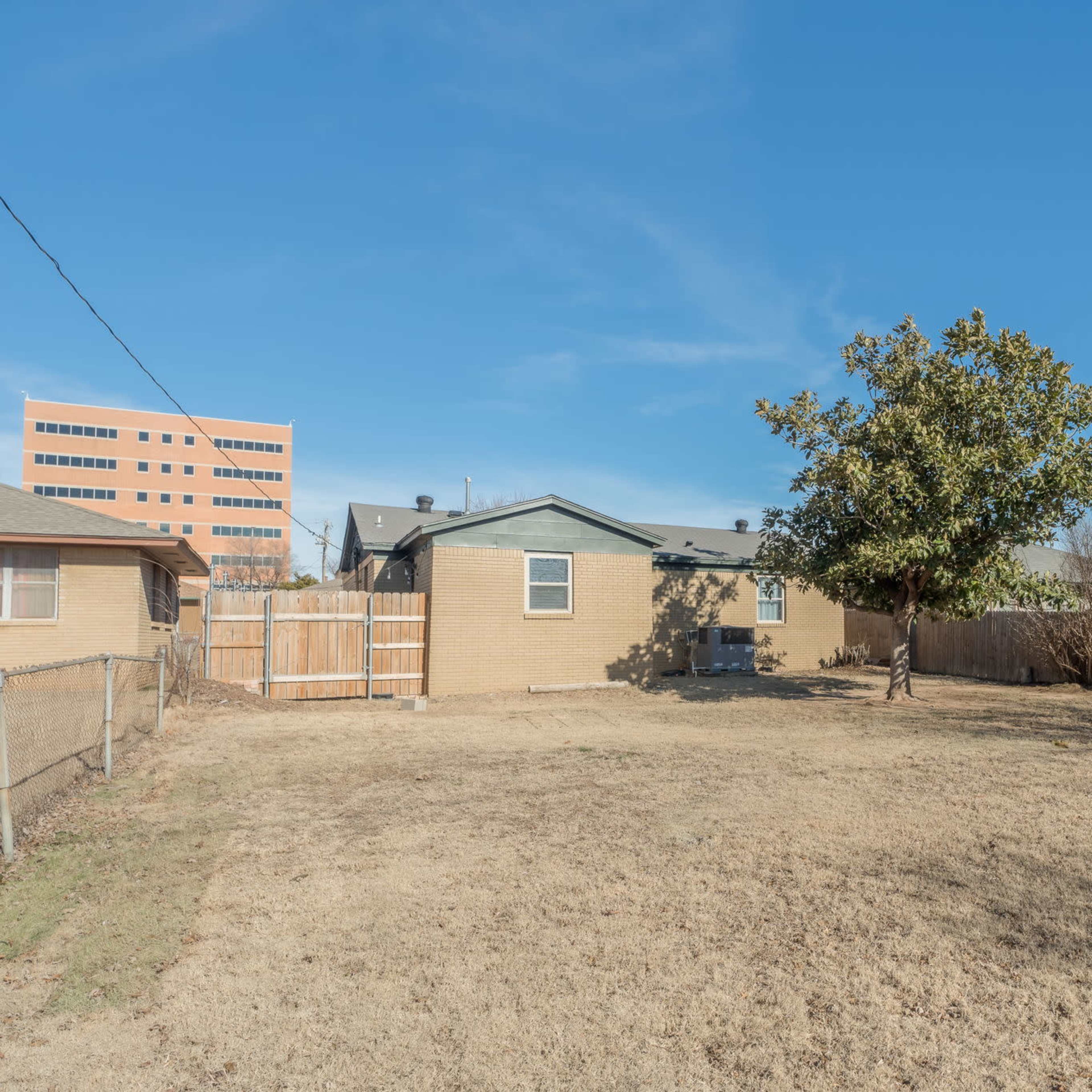 A dry backyard with a fenced area, a small tree, and a house in the background, alongside a larger building visible in the distance.