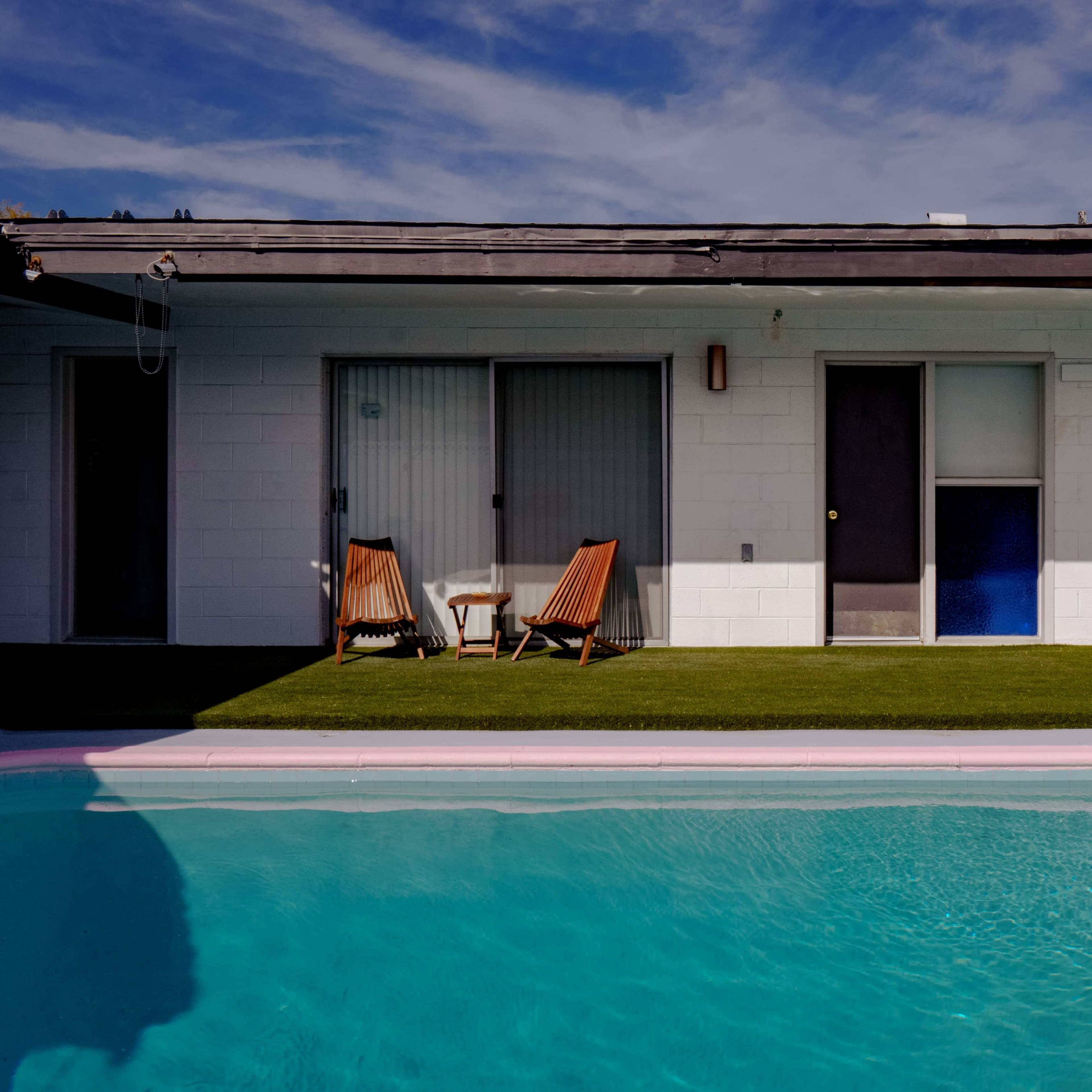 A clear blue swimming pool reflects a single-story building with multiple doors and two lounge chairs on a grassy area beside it.