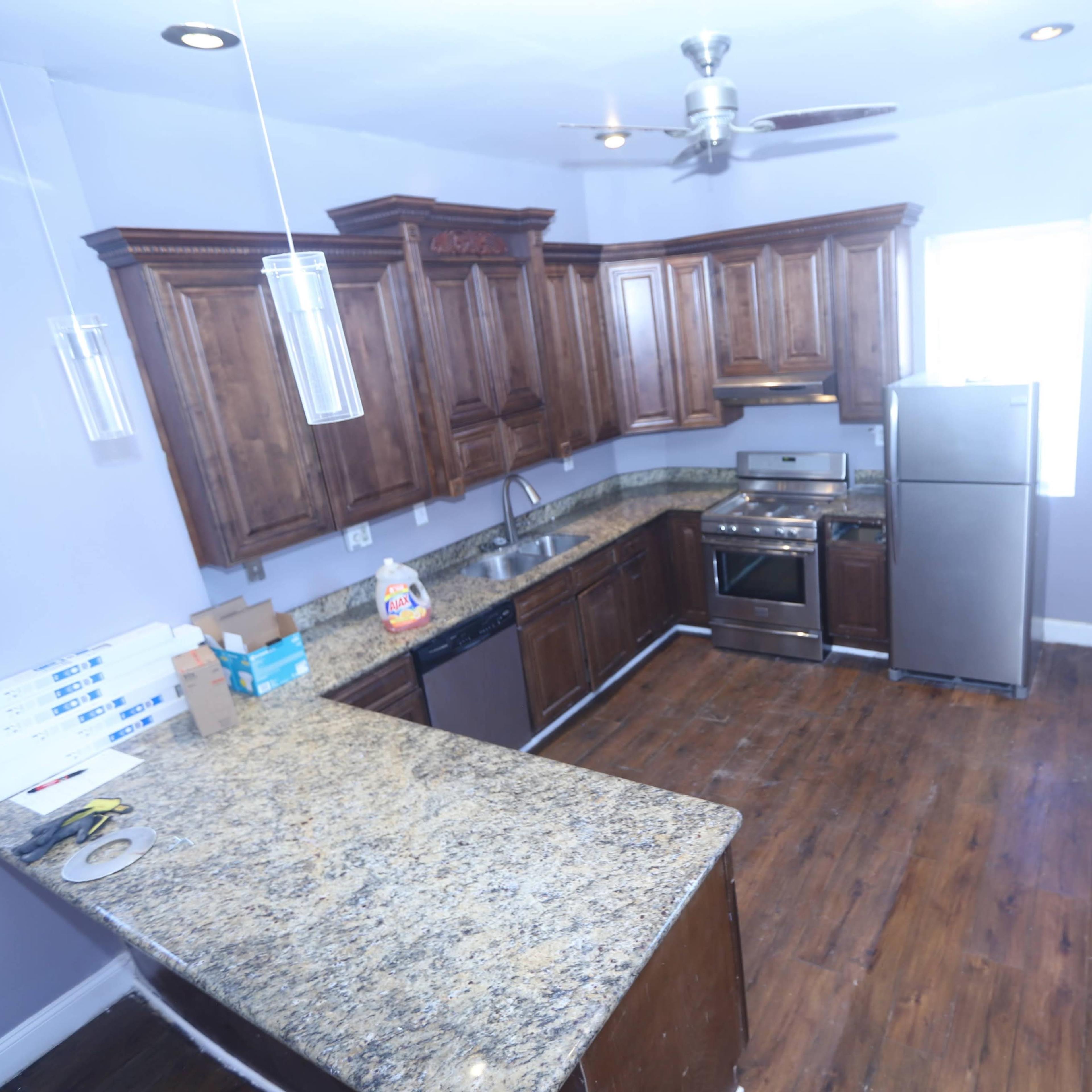 A kitchen with dark wooden cabinets, granite countertops, a stainless steel refrigerator, and an oven, featuring a wooden floor and a ceiling fan.