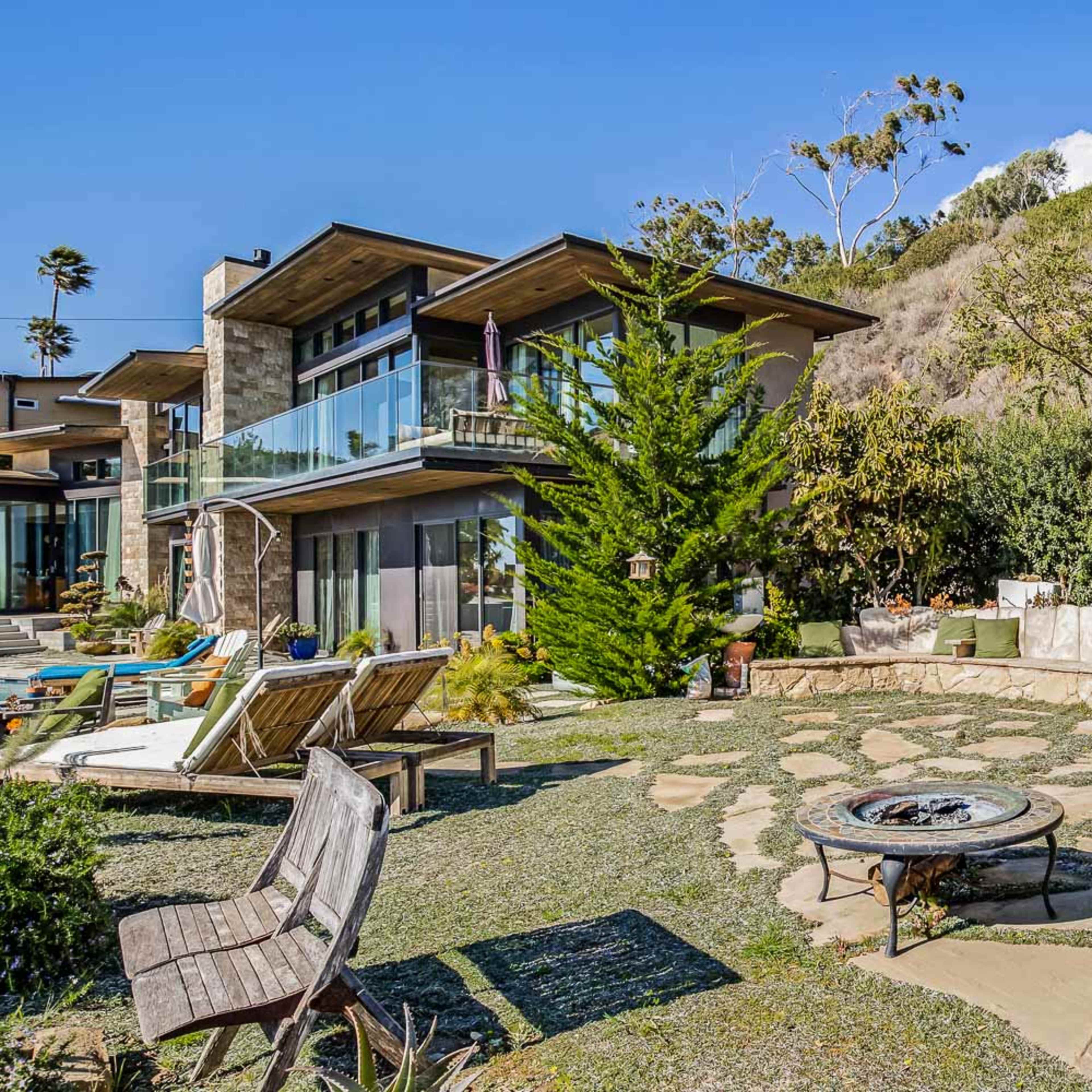 A modern house surrounded by landscaped greenery and a stone pathway leading to a circular seating area with a fire pit.