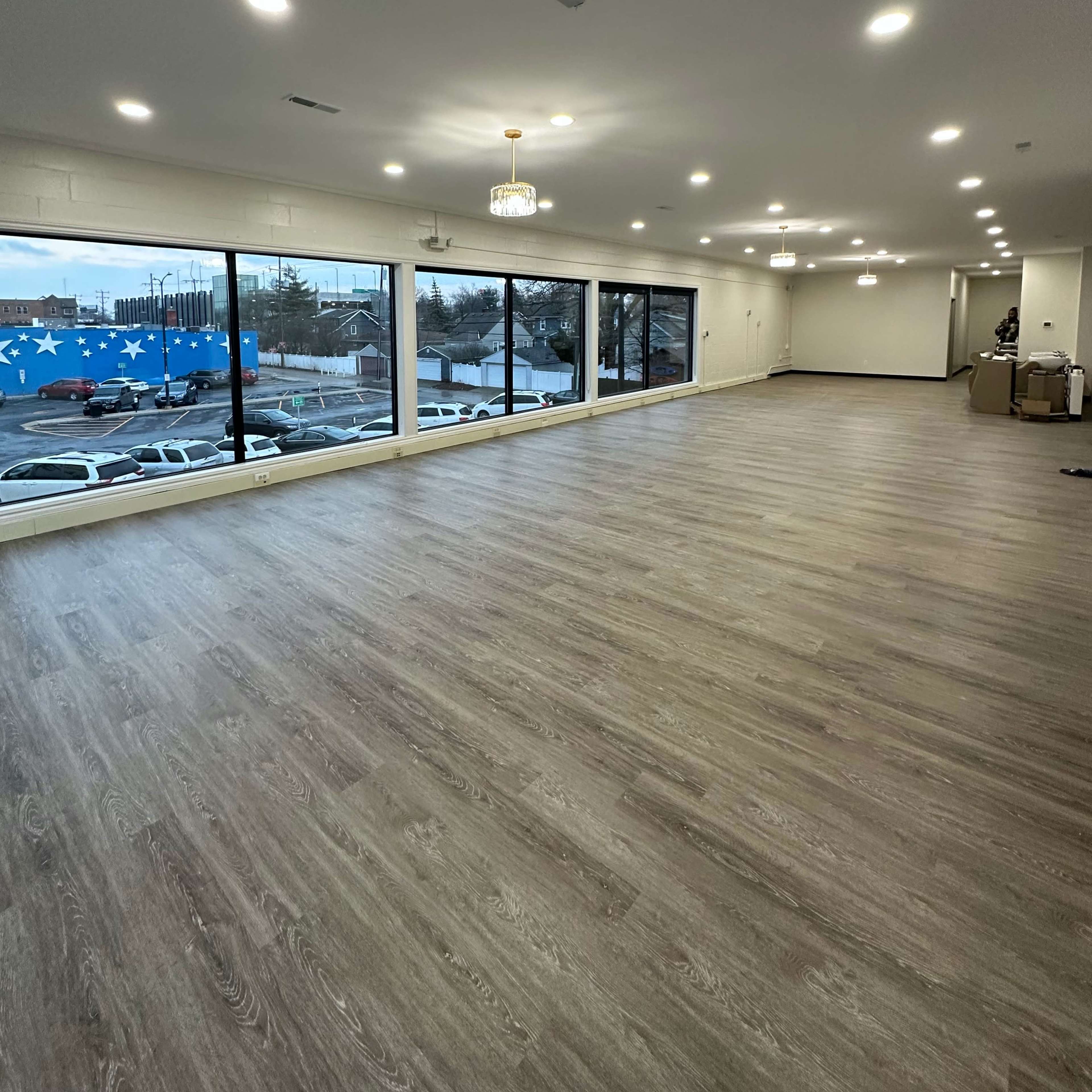 A spacious, empty room with wooden flooring and large windows overlooking a parking lot.