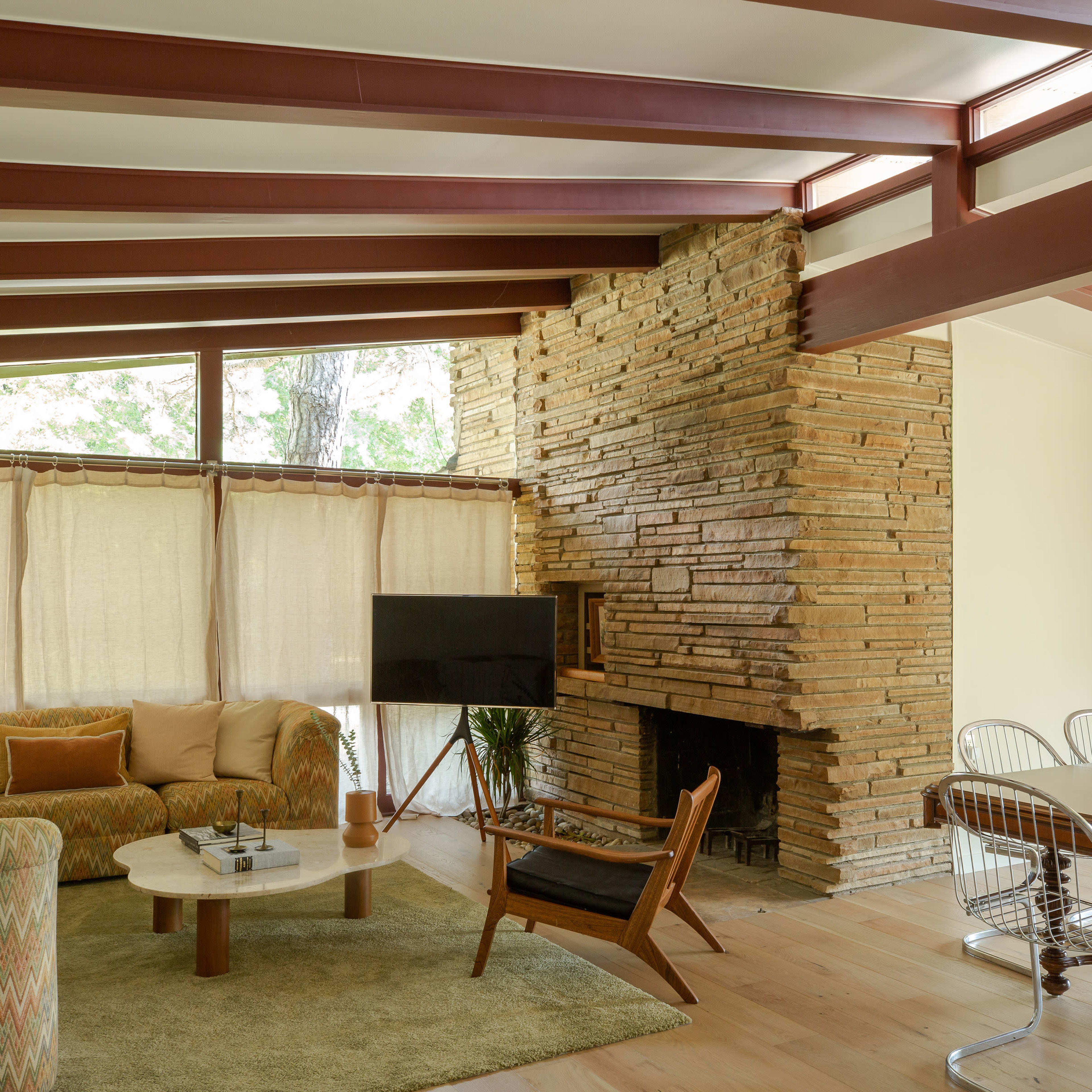 A mid-century modern living room featuring a stone fireplace, a circular coffee table, a beige sofa, and a dining area with a wooden table and metal chairs.