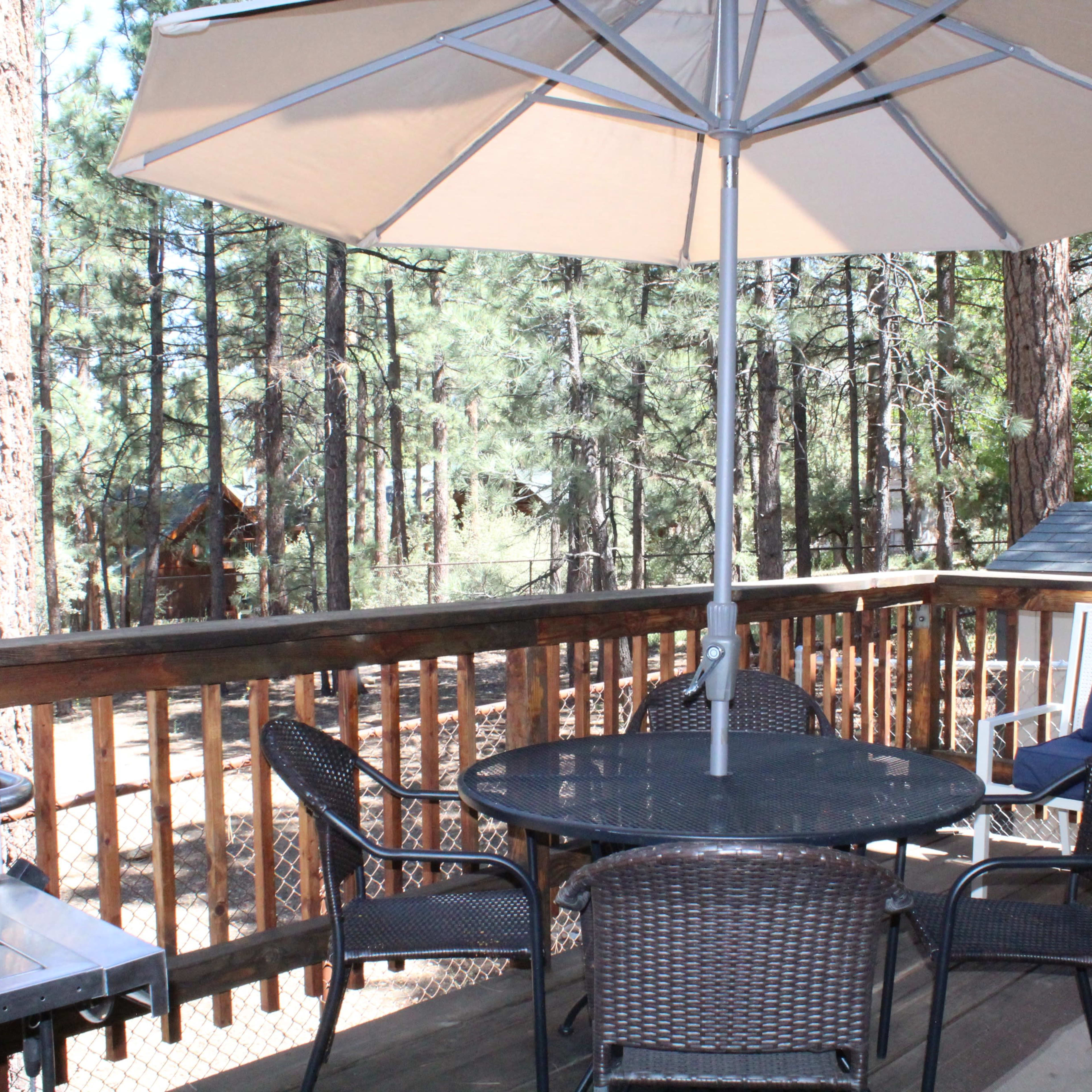 A round table with chairs and a large umbrella is set on a wooden deck surrounded by tall trees.