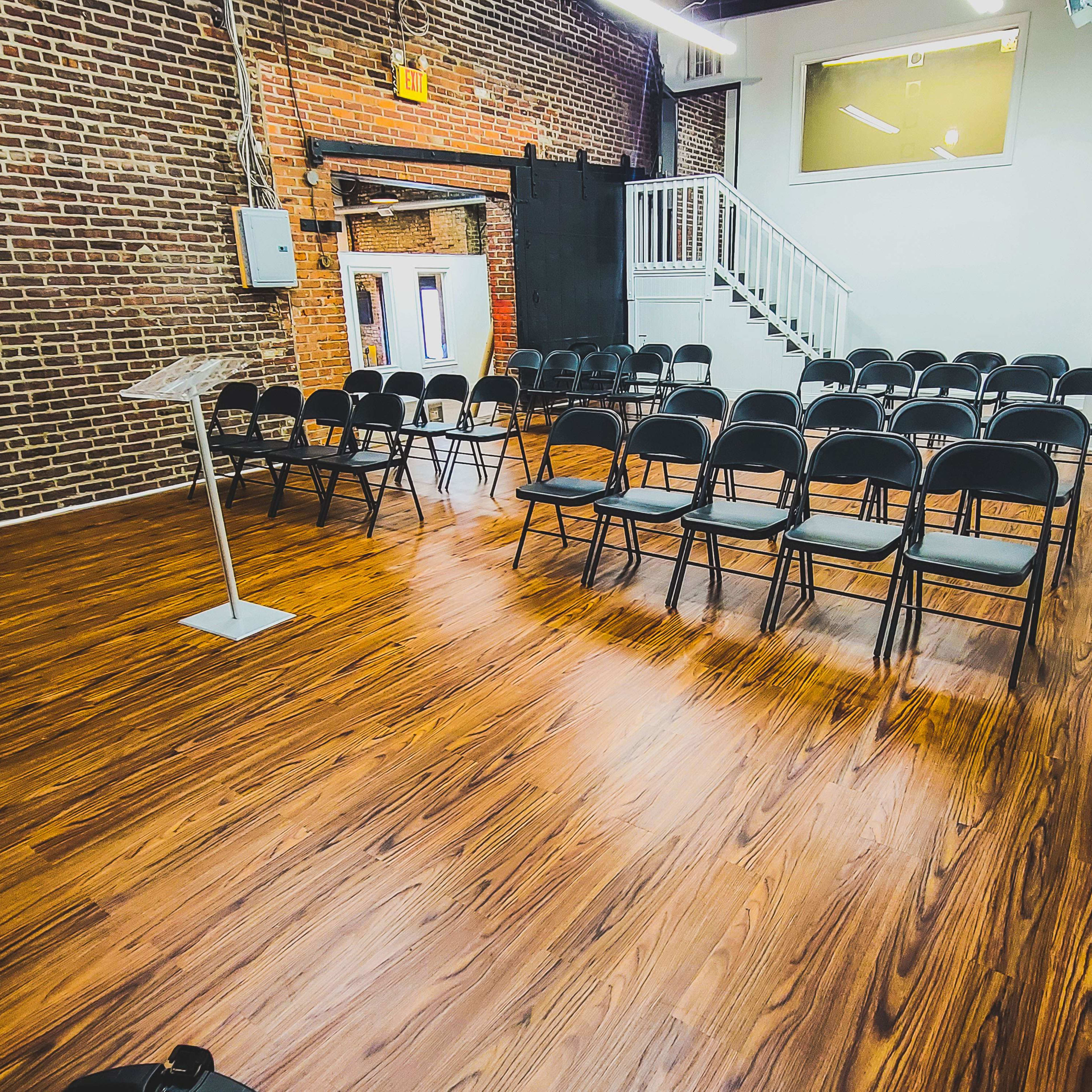A room with a wooden floor features rows of black chairs facing a clear lectern, against a backdrop of exposed brick walls.