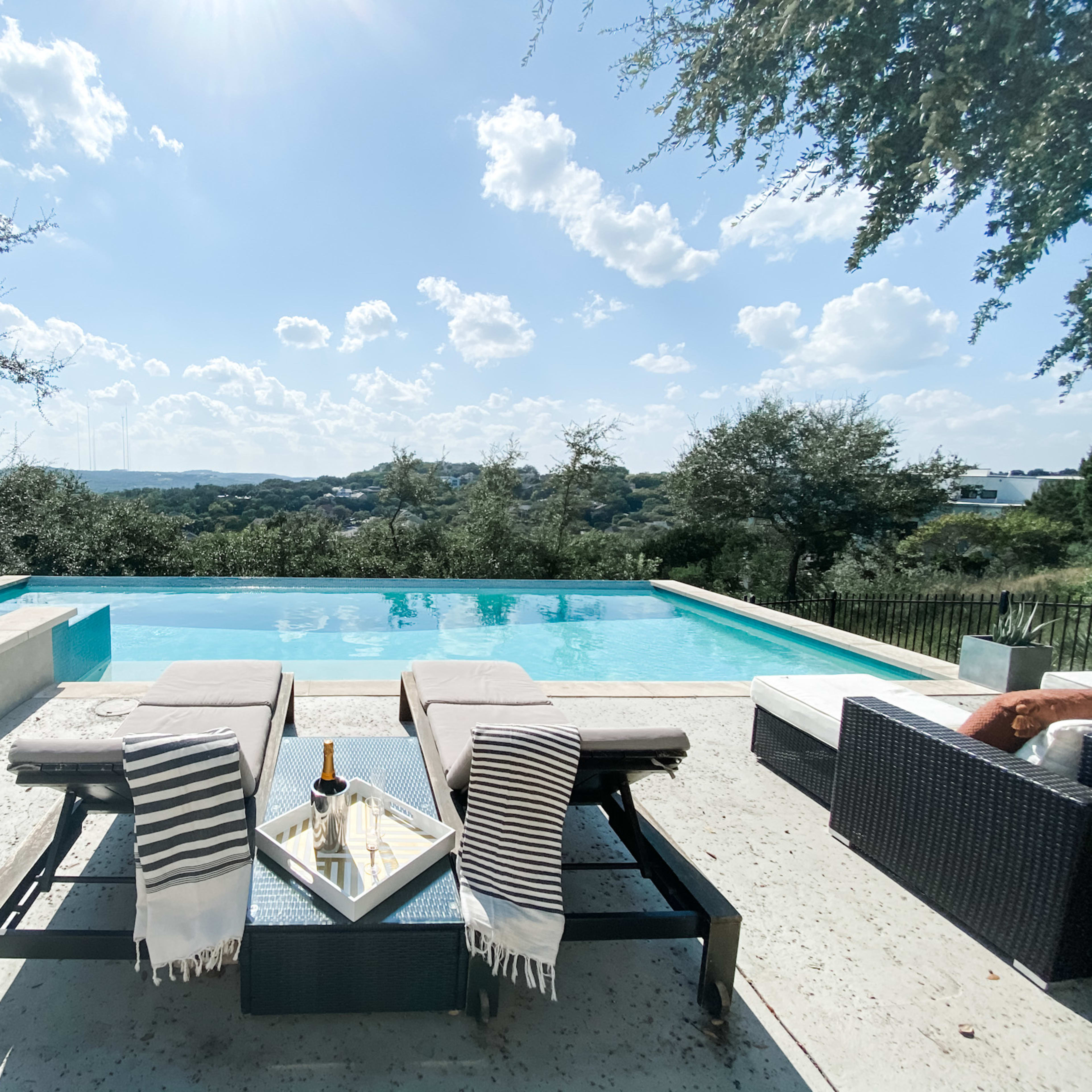 A clear swimming pool with lounge chairs and a table is set against a backdrop of rolling hills and a blue sky.