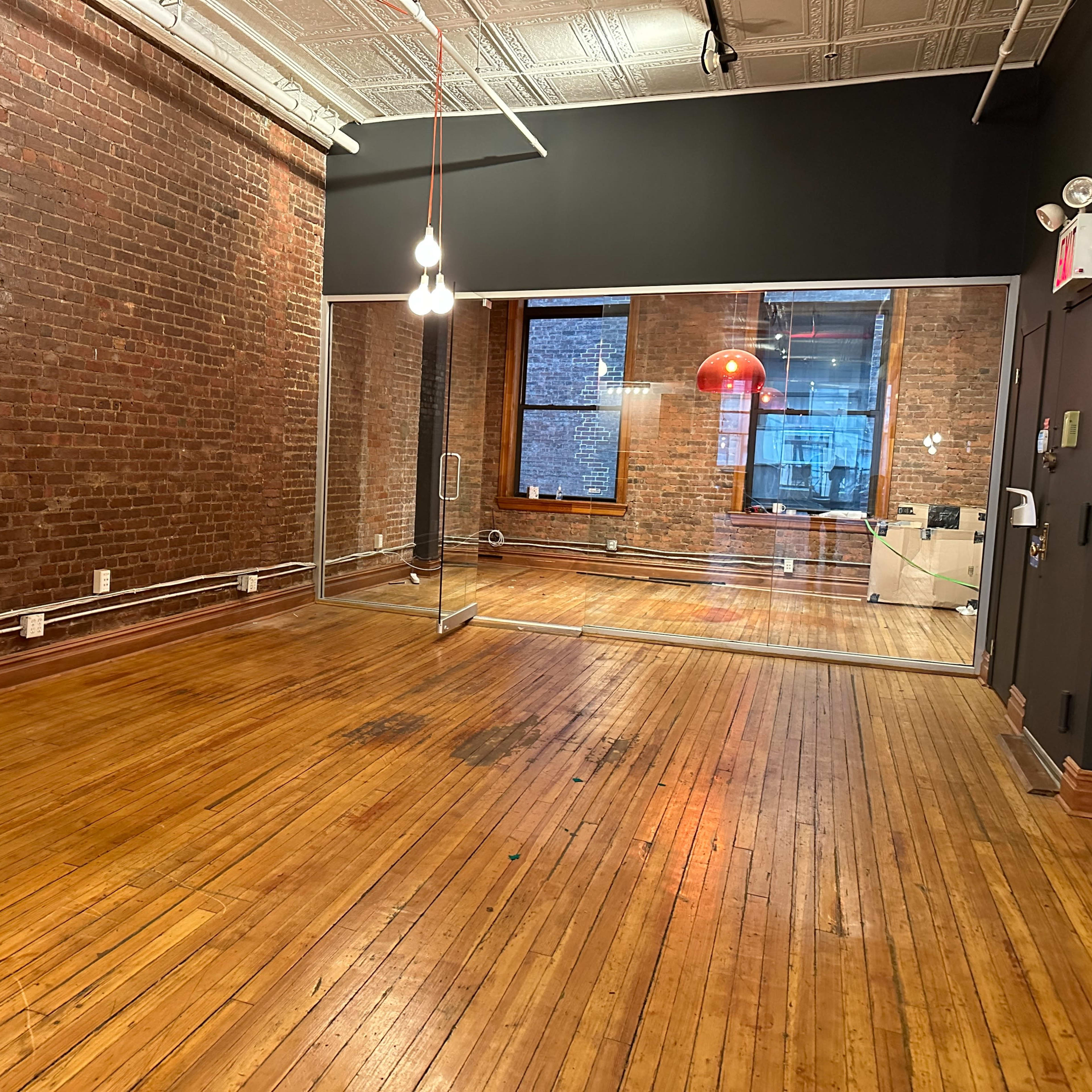 A spacious room with exposed brick walls, wooden flooring, and a large glass wall separating a smaller room with a red pendant light.