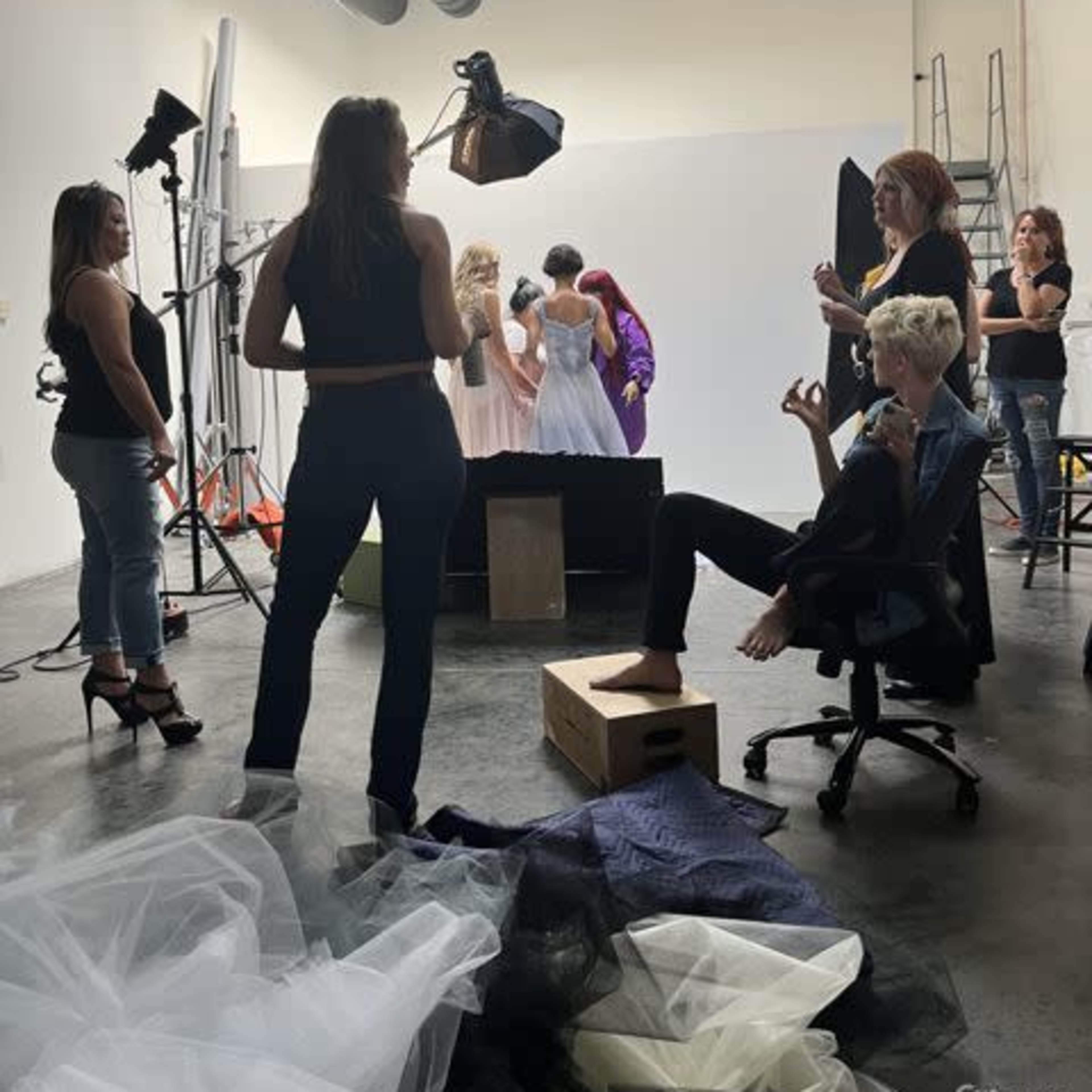 A group of people works in a studio setting, with some preparing for a photo shoot while others pose in elegant dresses.