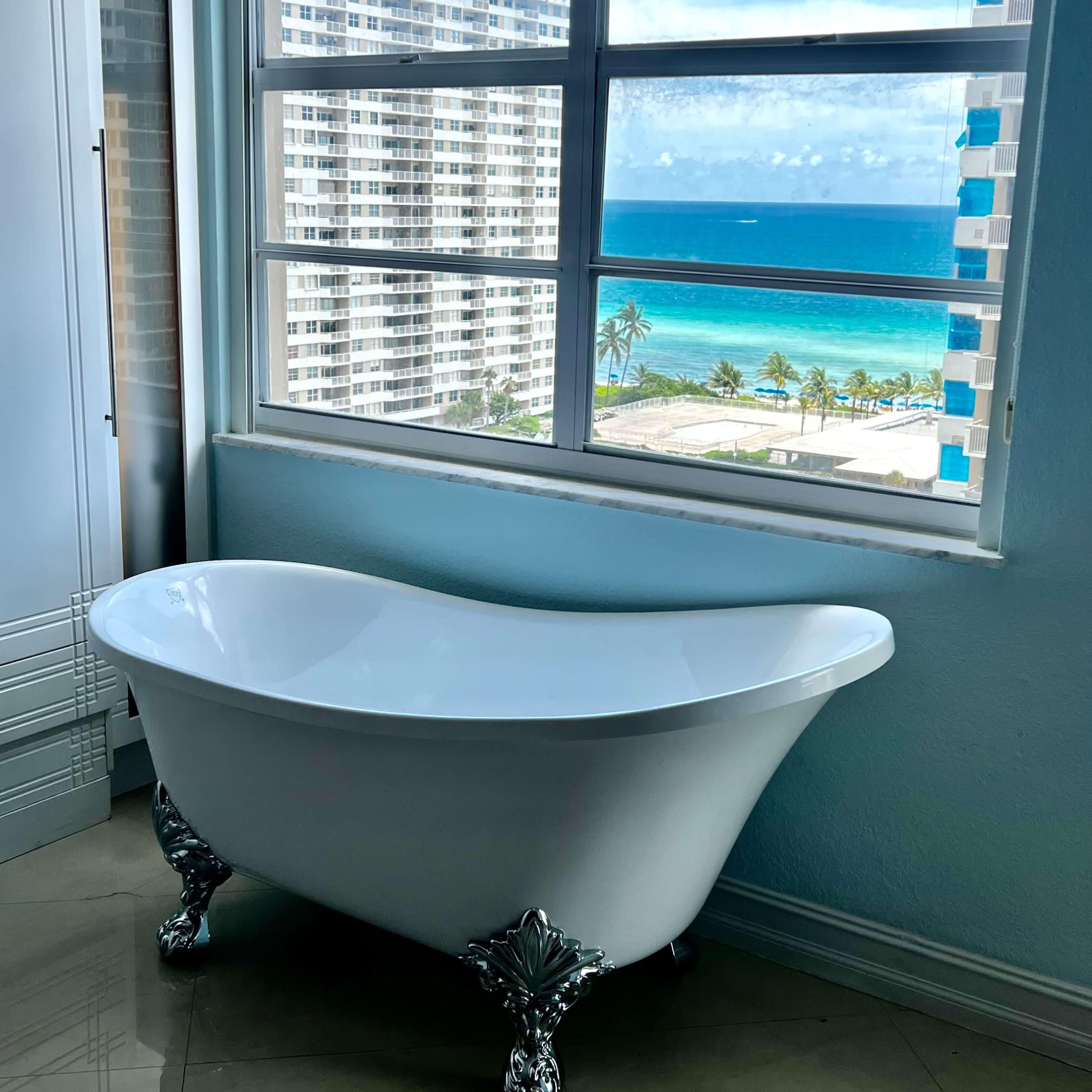 A freestanding white bathtub with chrome feet is positioned near a large window overlooking a view of the ocean and buildings.