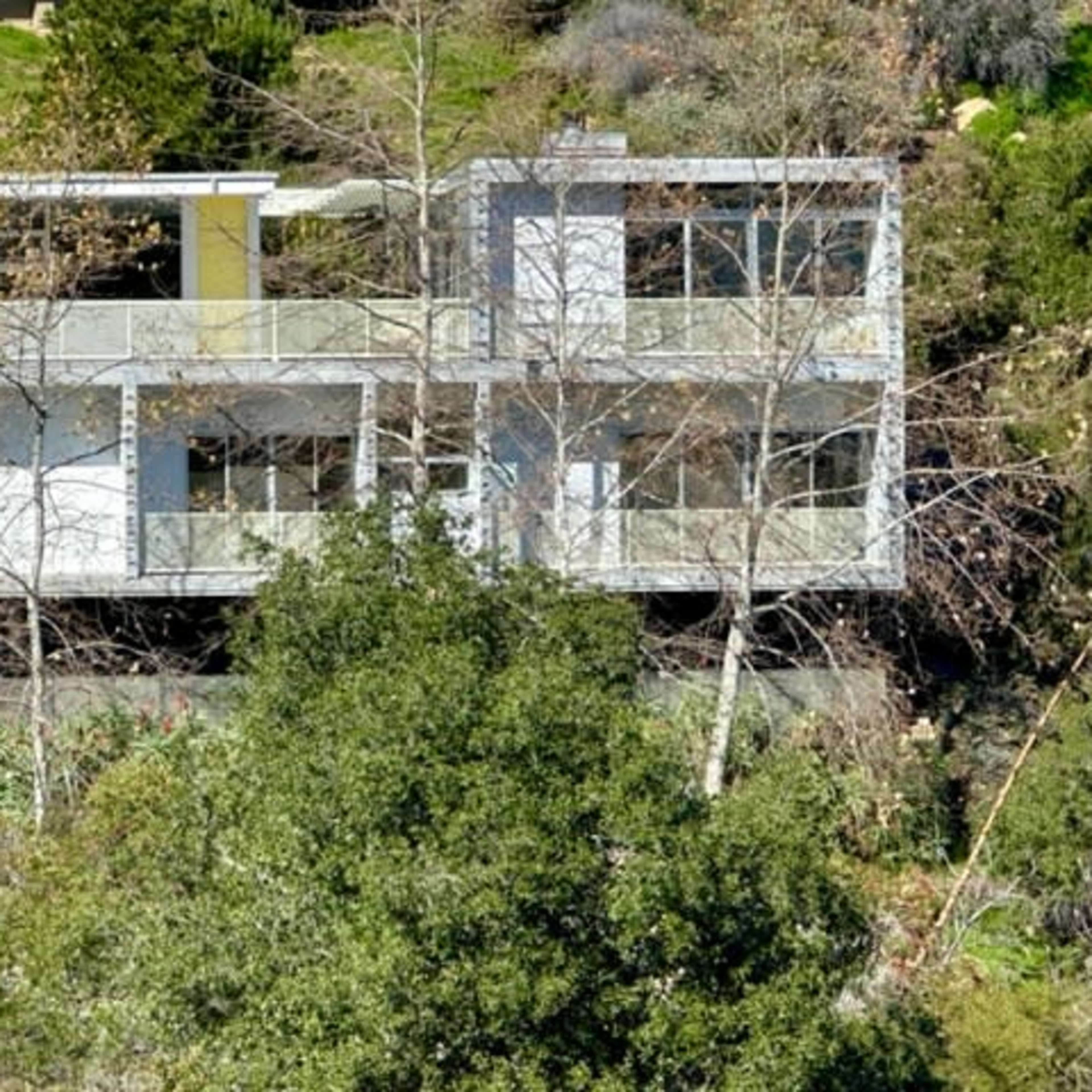 A modern two-story house with large windows and balconies, situated on a hillside among greenery.