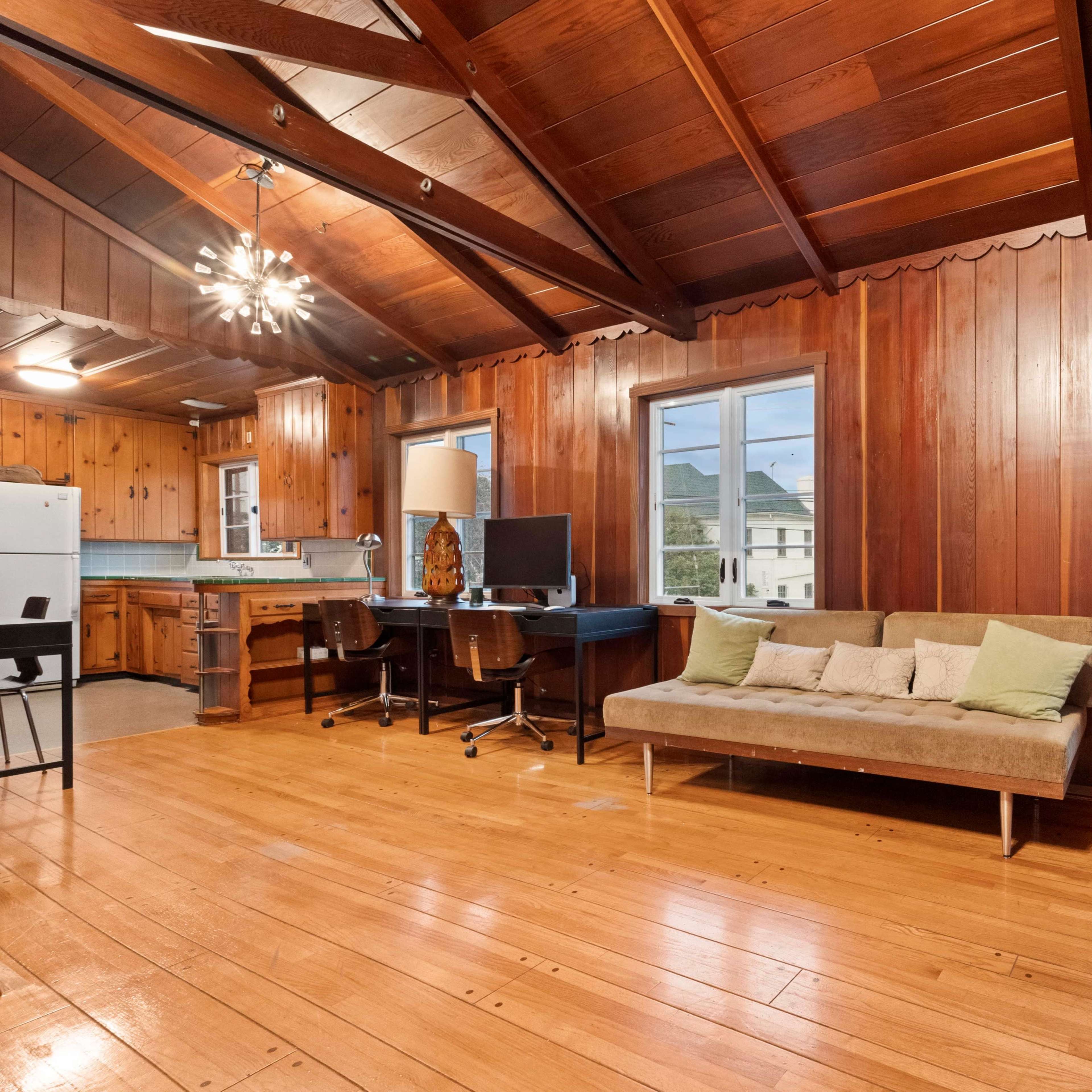 The room features a wooden interior with a sofa, desk, and computer, surrounded by large windows that allow natural light in.