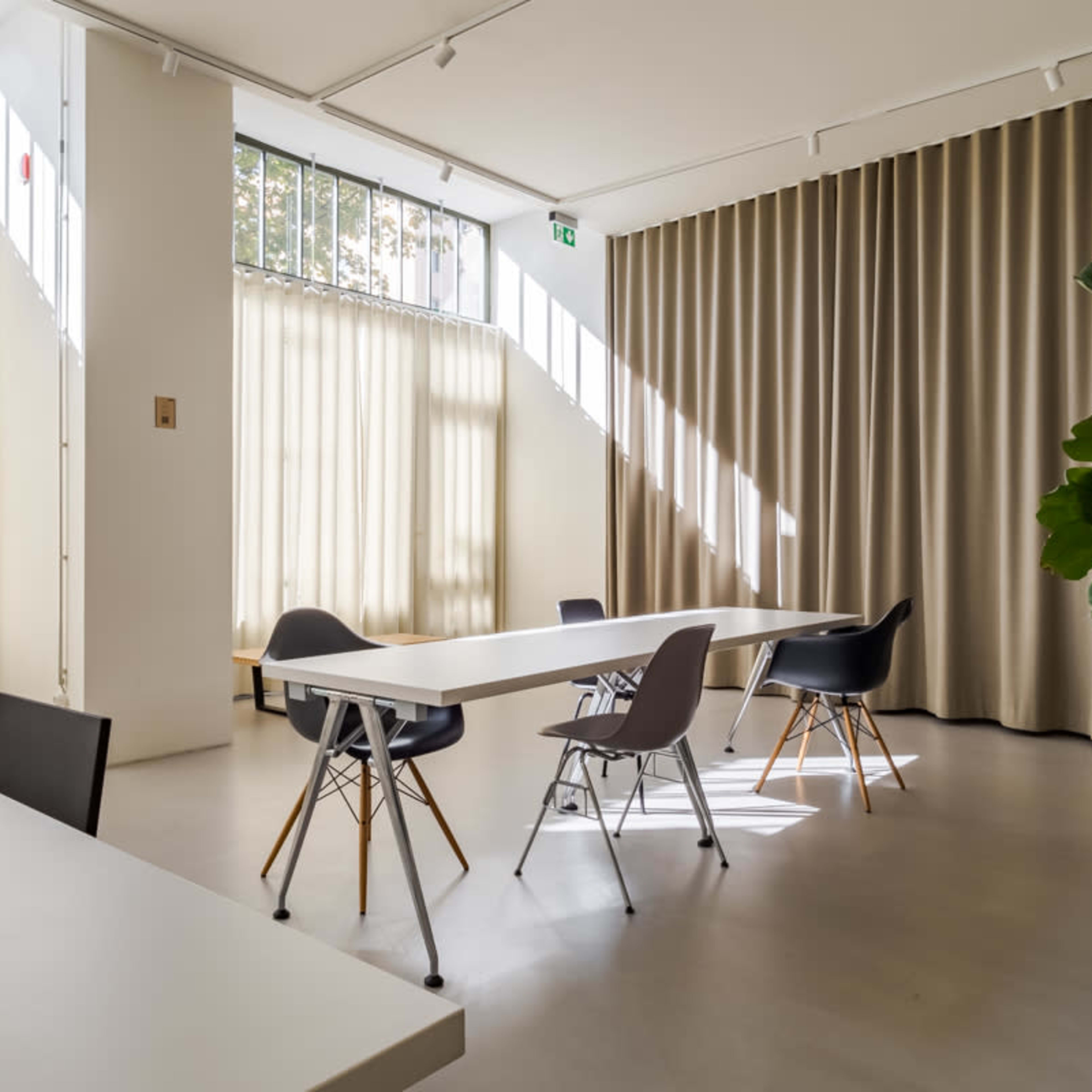 A modern, minimalist room with a long white table, several chairs, large windows with curtains, and a potted plant in the corner.