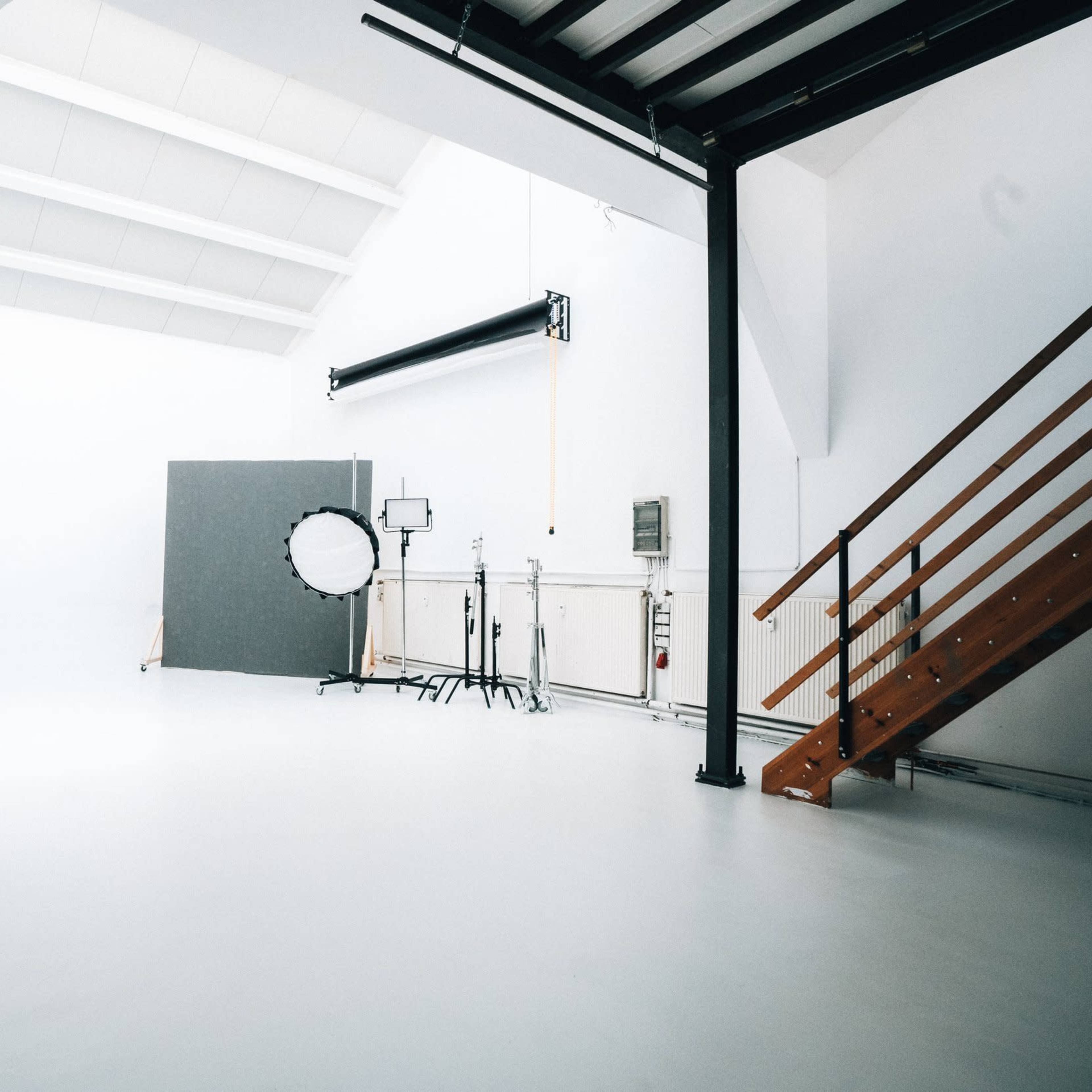 A well-lit photography studio with a white background, a large round reflector on a stand, and a wooden staircase leading to a lofted area.