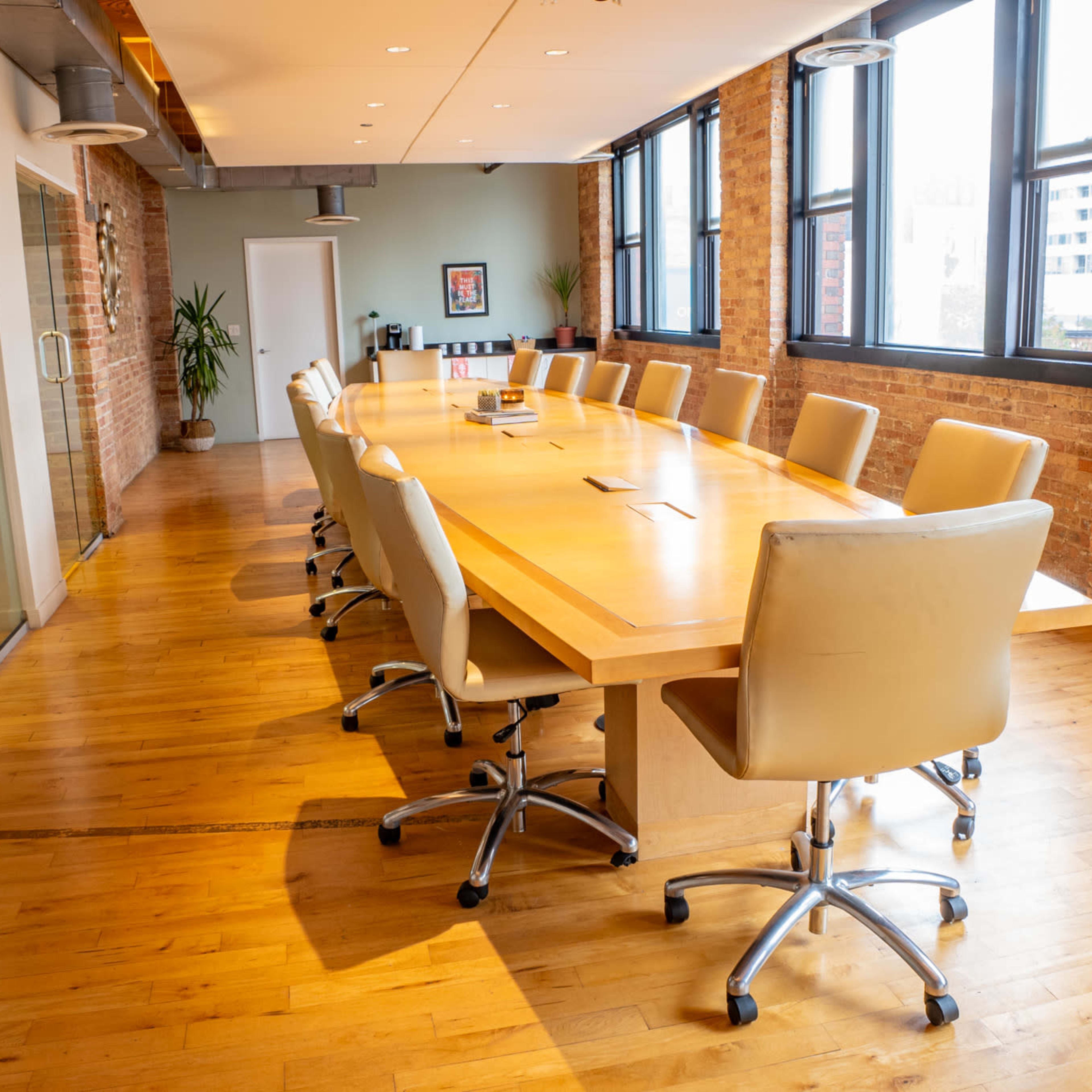 A spacious conference room features a long wooden table surrounded by leather chairs, with large windows allowing natural light to illuminate the space.